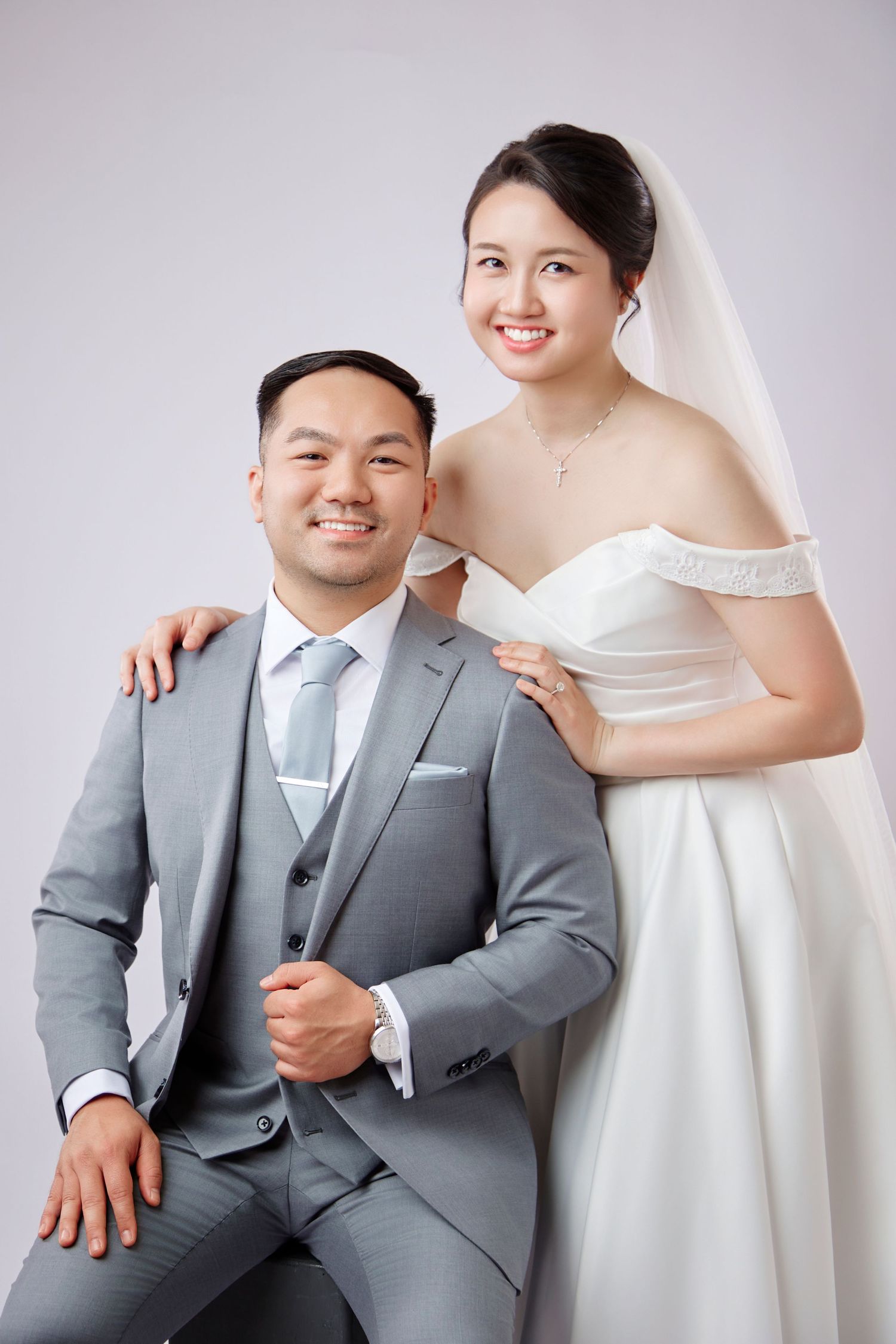 Smiling Asian bride in white off-shoulder gown and veil poses with groom in gray suit.