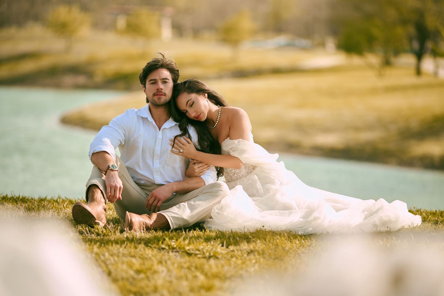 Romantic couple sitting on grass by lake in golden sunset light, bride in white gown.
