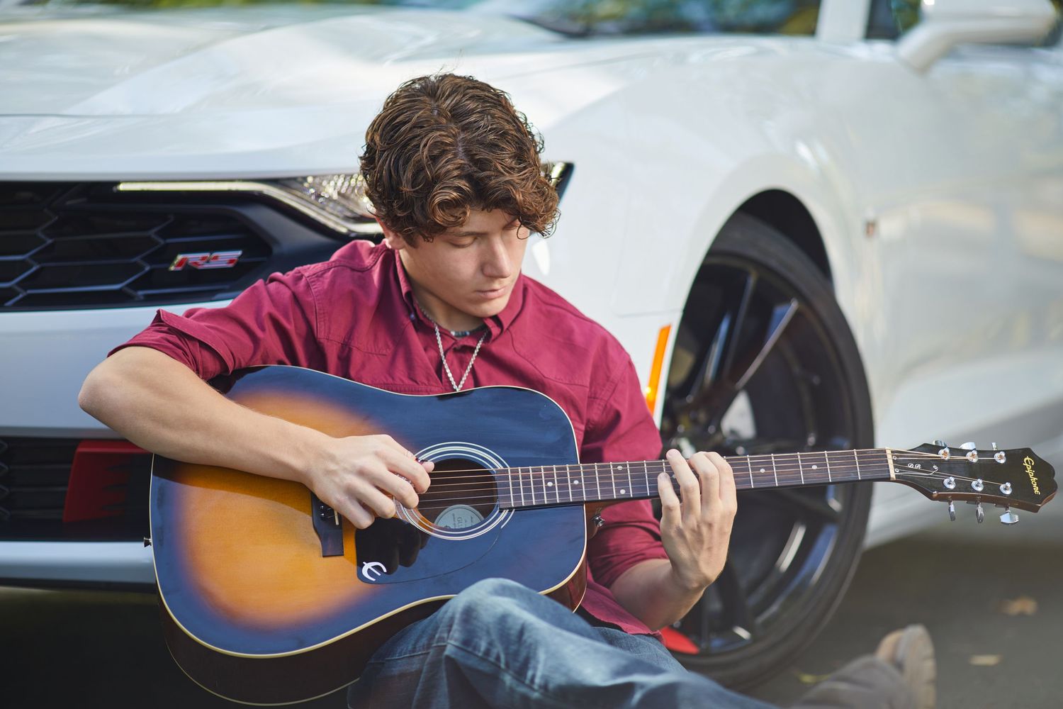 Young man in red shirt playing acoustic guitar while sitting beside a white sports car outdoors.