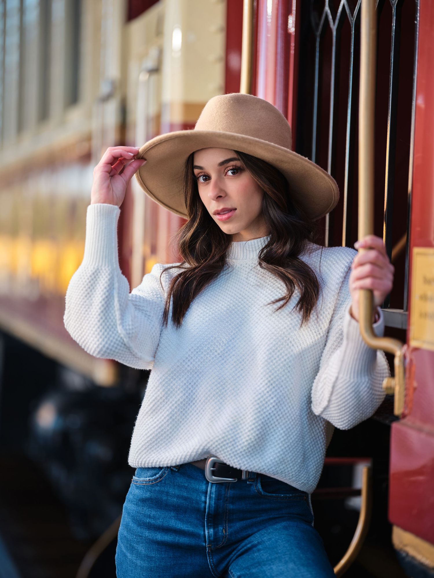 Brunette woman in white sweater and wide-brim hat posing beside a vintage red train car.