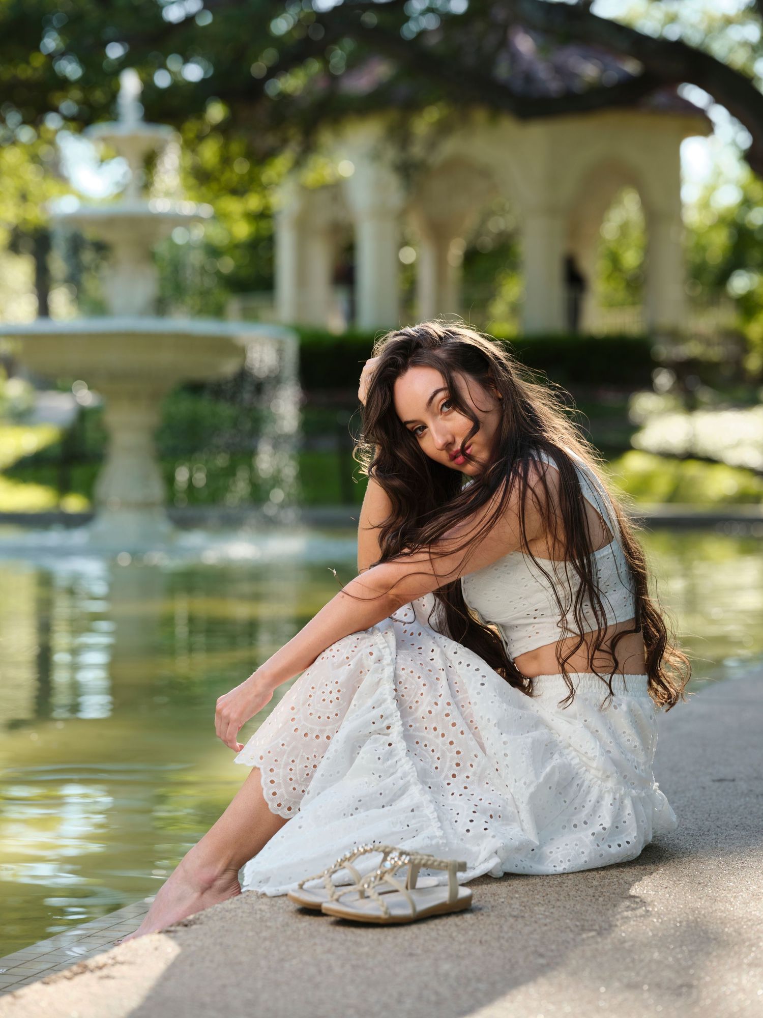 Woman in white eyelet skirt sitting by a garden fountain in golden sunlight.