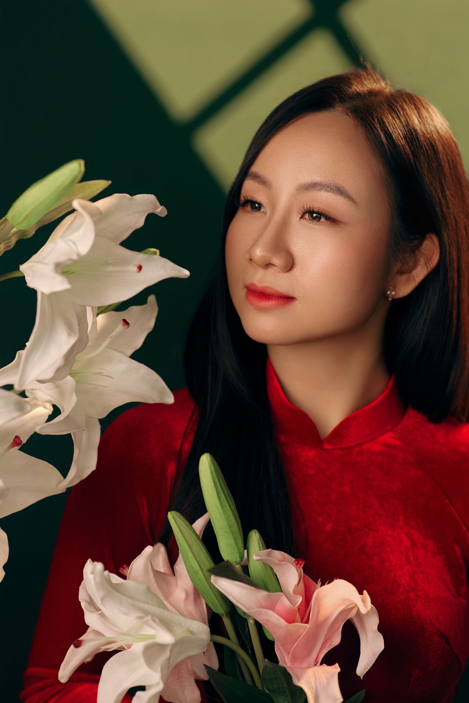 Elegant Asian woman in red ao dai holds white and pink lilies in green-lit studio.