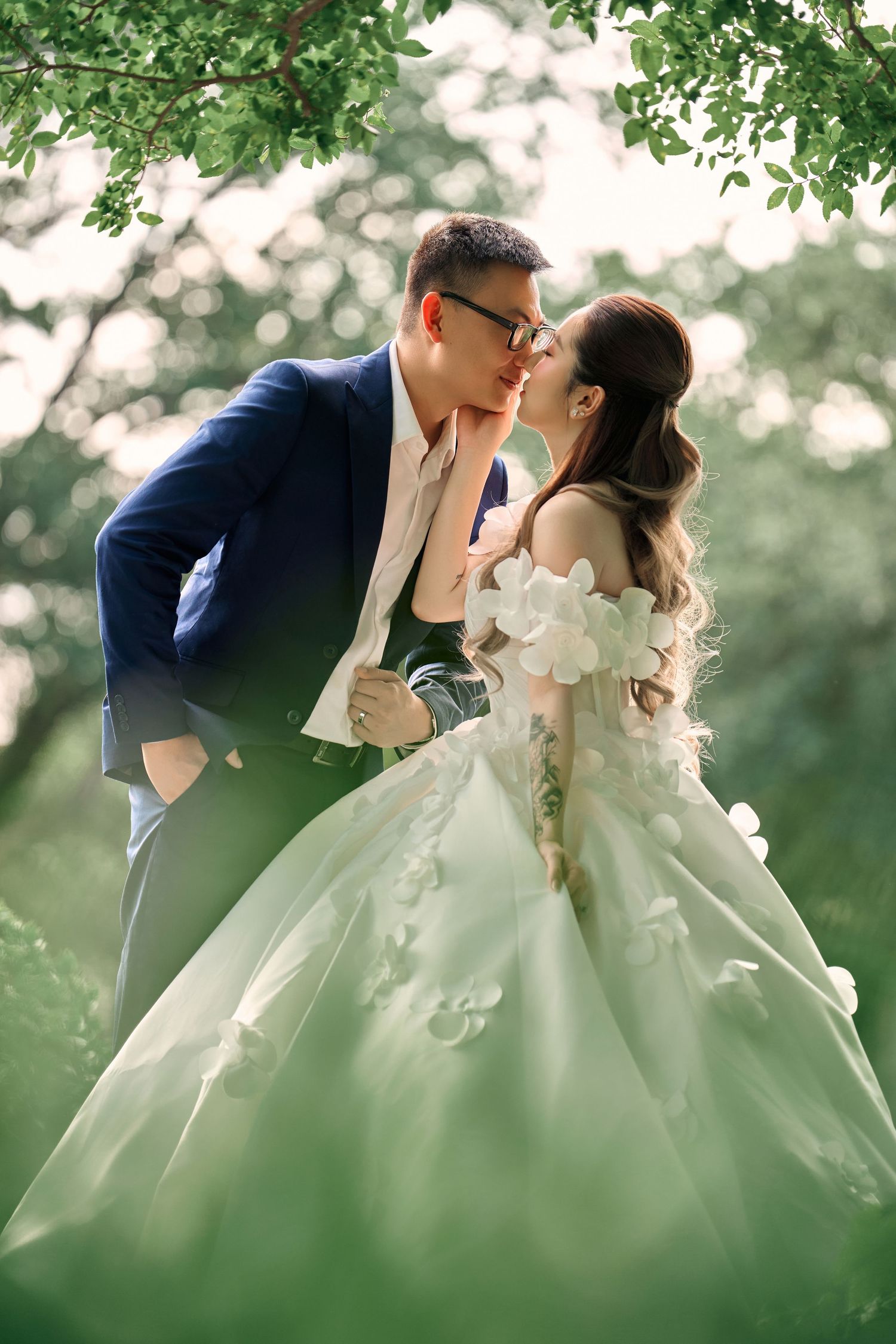 A couple shares a romantic kiss outdoors, bride in a floral green gown and groom in a navy suit, surrounded by lush greenery.
