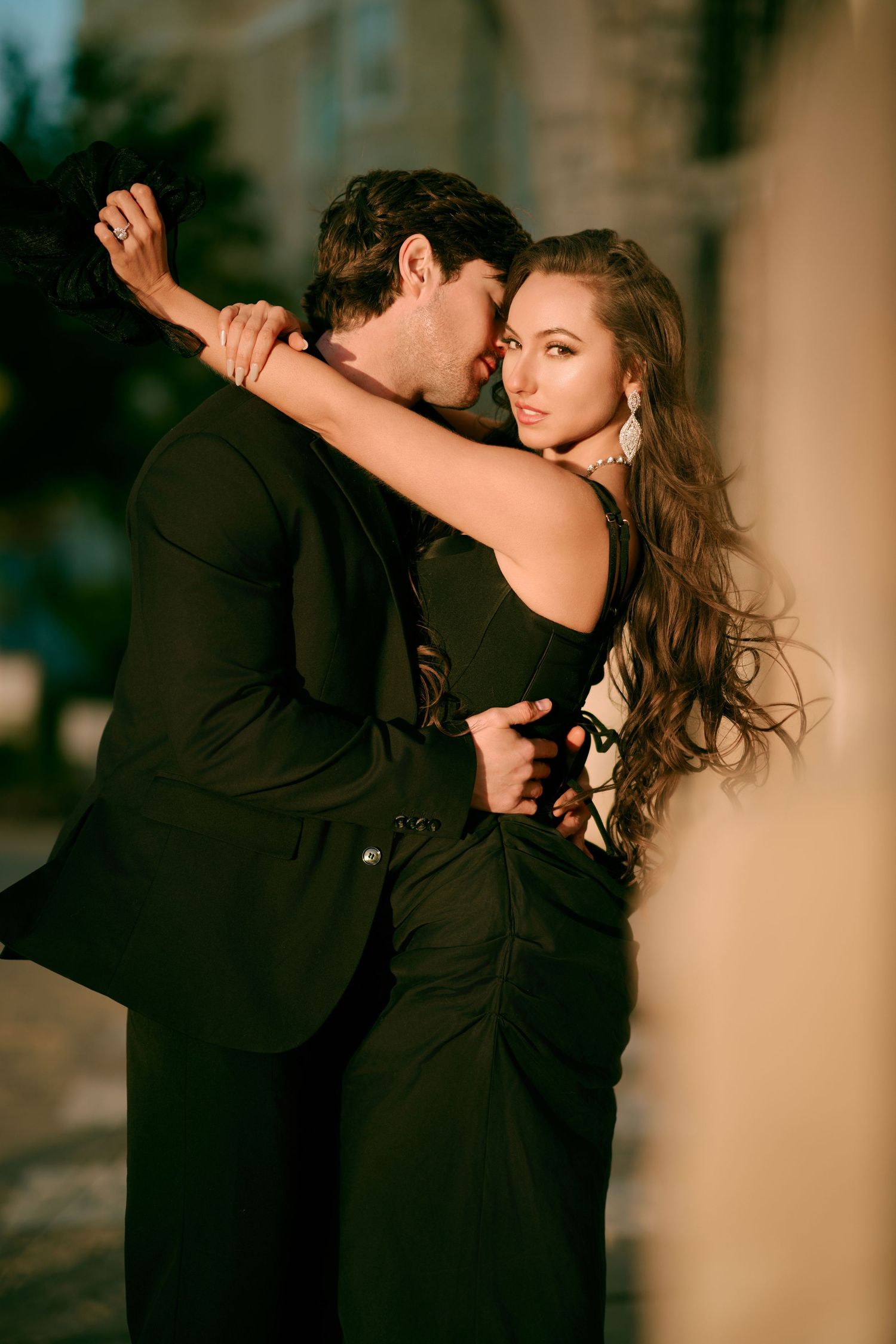 Elegant couple in black outfits embracing romantically on a city street at golden hour.
