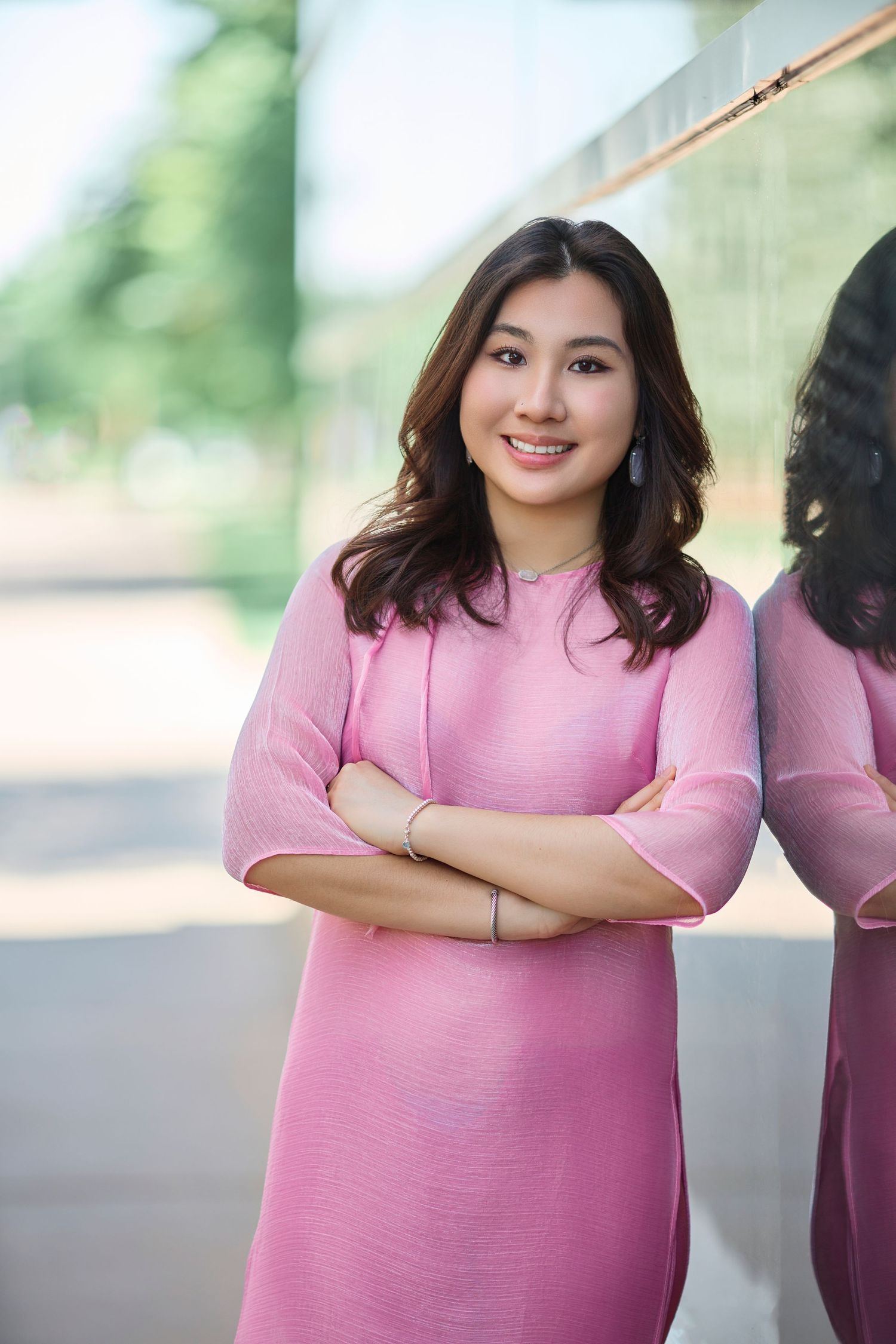 Smiling young Asian woman in pink dress standing with arms crossed near reflective glass wall.