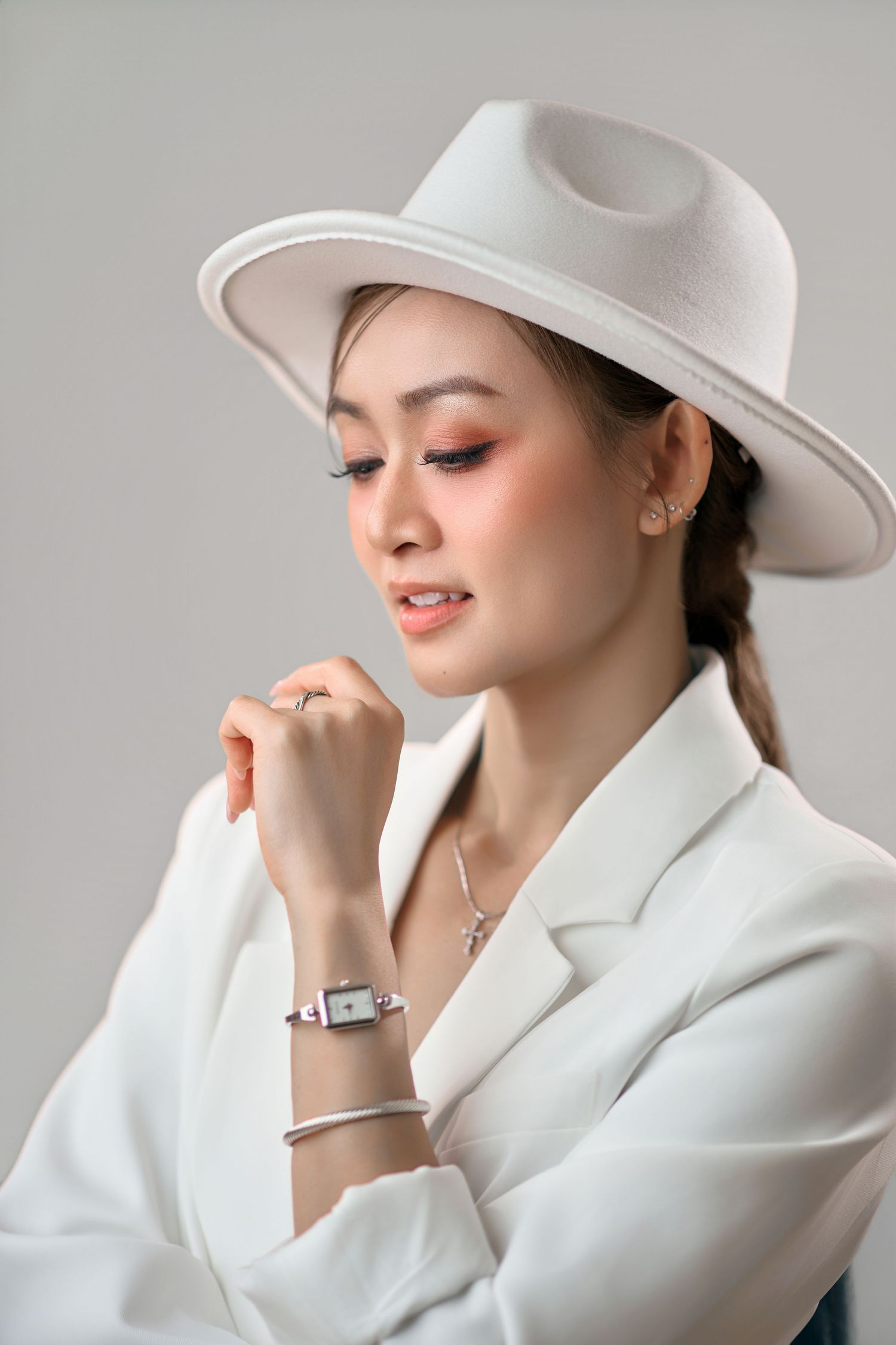 Elegant Asian woman in white blazer and hat looking down with wrist accessories on gray background.