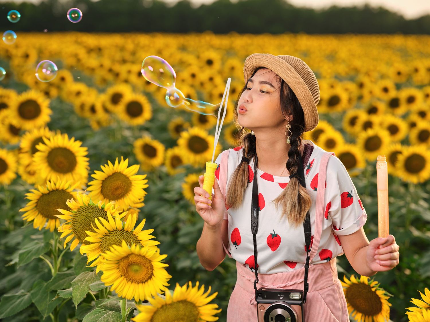 Young woman in straw hat blowing bubbles with camera strap in a vibrant sunflower field.