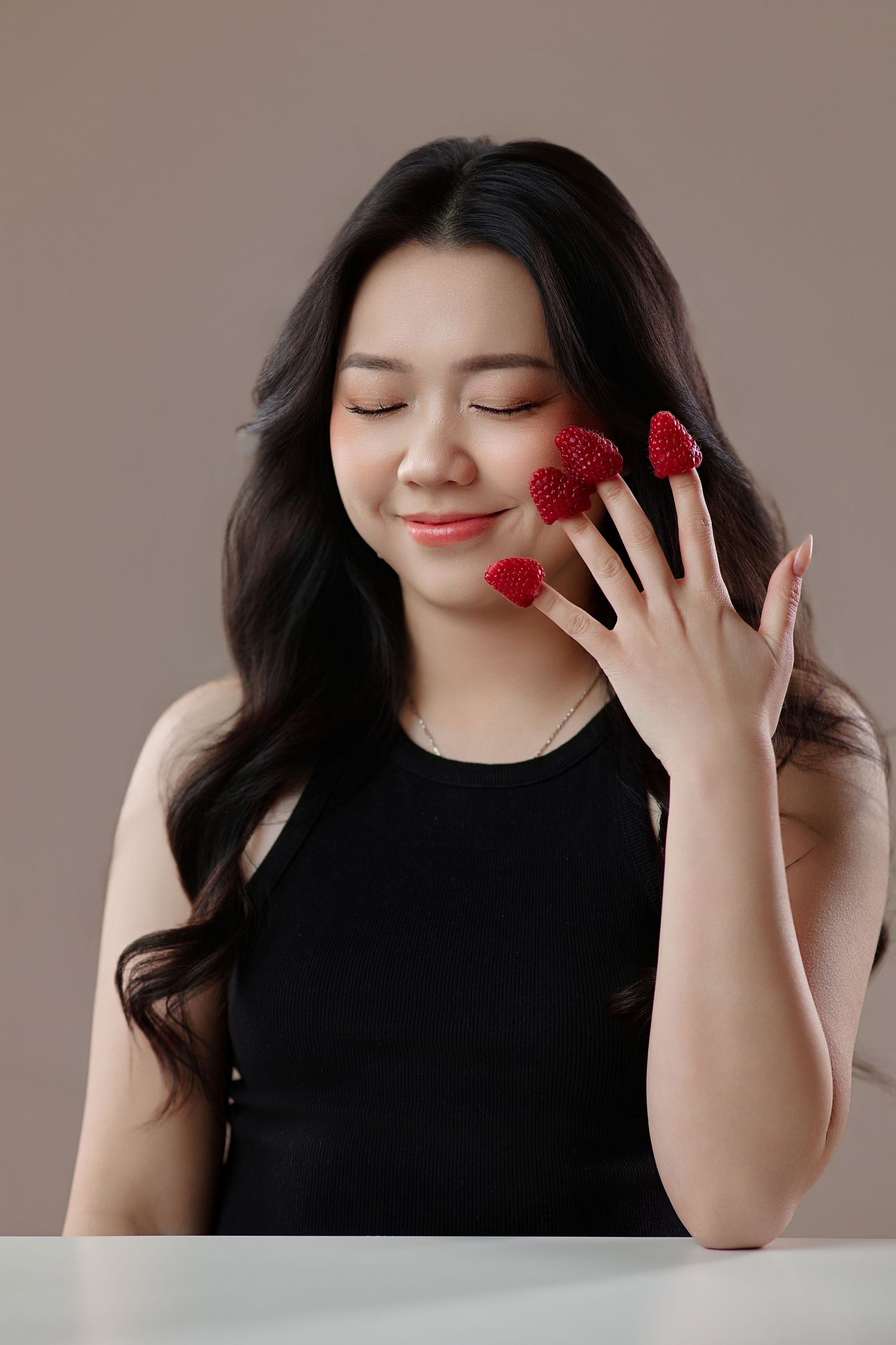 Asian woman with raspberries on fingers smiling with closed eyes in black top.