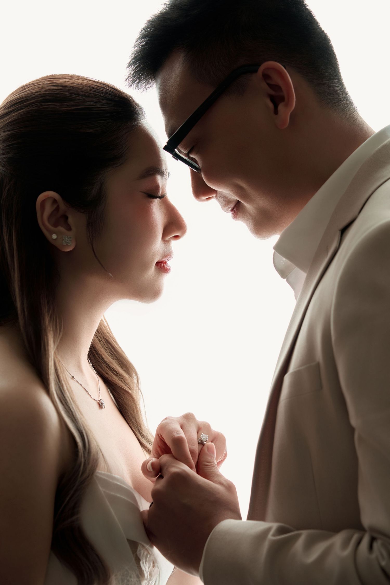A couple holds hands intimately, the man in glasses and suit gazing down at the woman in a tender romantic moment.