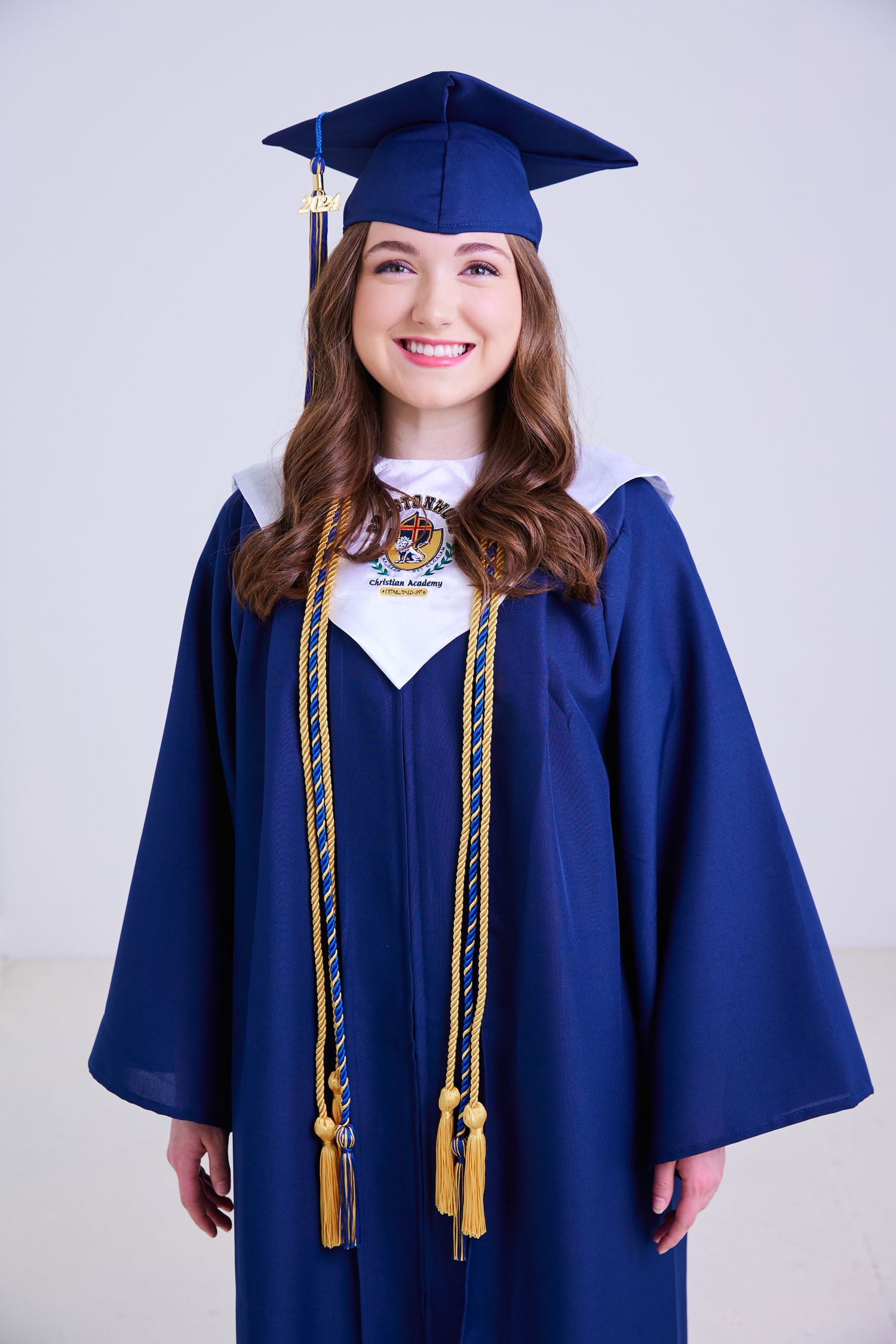 Smiling female graduate in navy blue cap and gown with gold honor cords on white background.