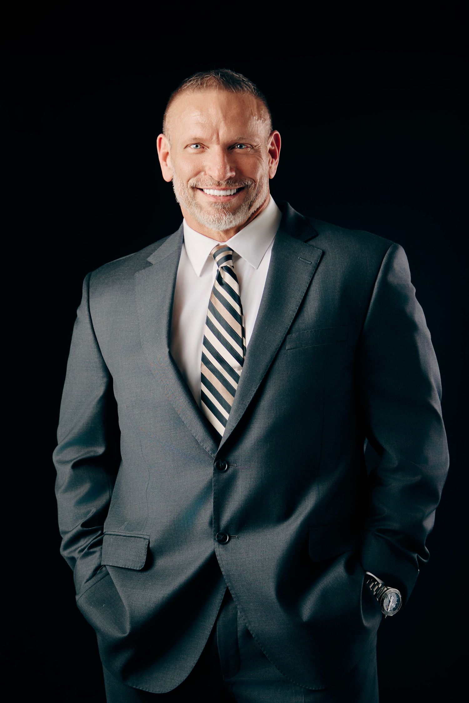 A confident businessman in a gray suit and striped tie smiles with hands in pockets against a black background.