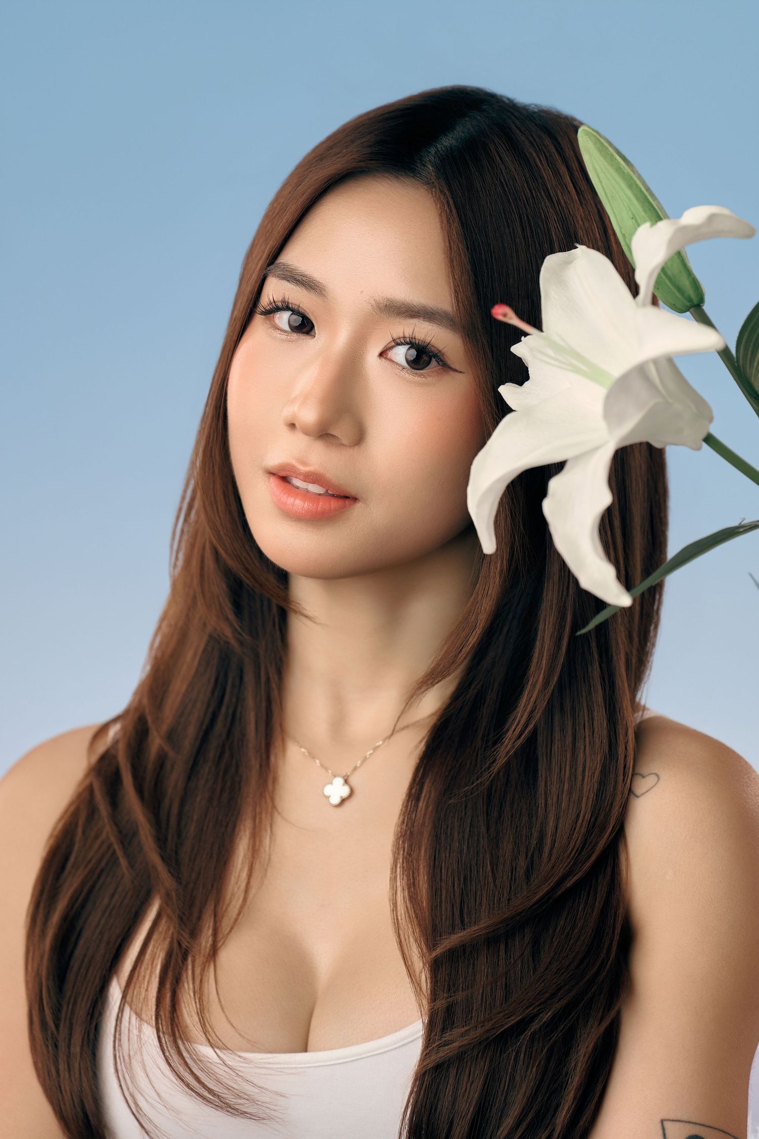 Young Asian woman with long brown hair poses with a white lily flower against a soft blue background.
