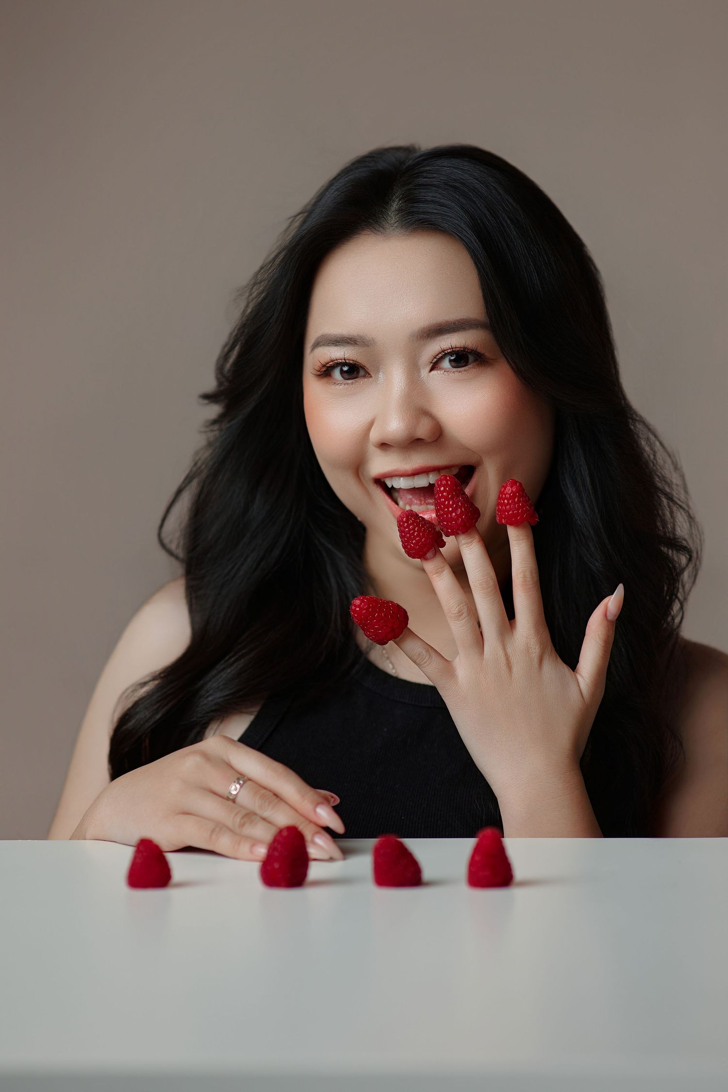 Smiling Asian woman holding raspberries on fingers near mouth with fruits on table.