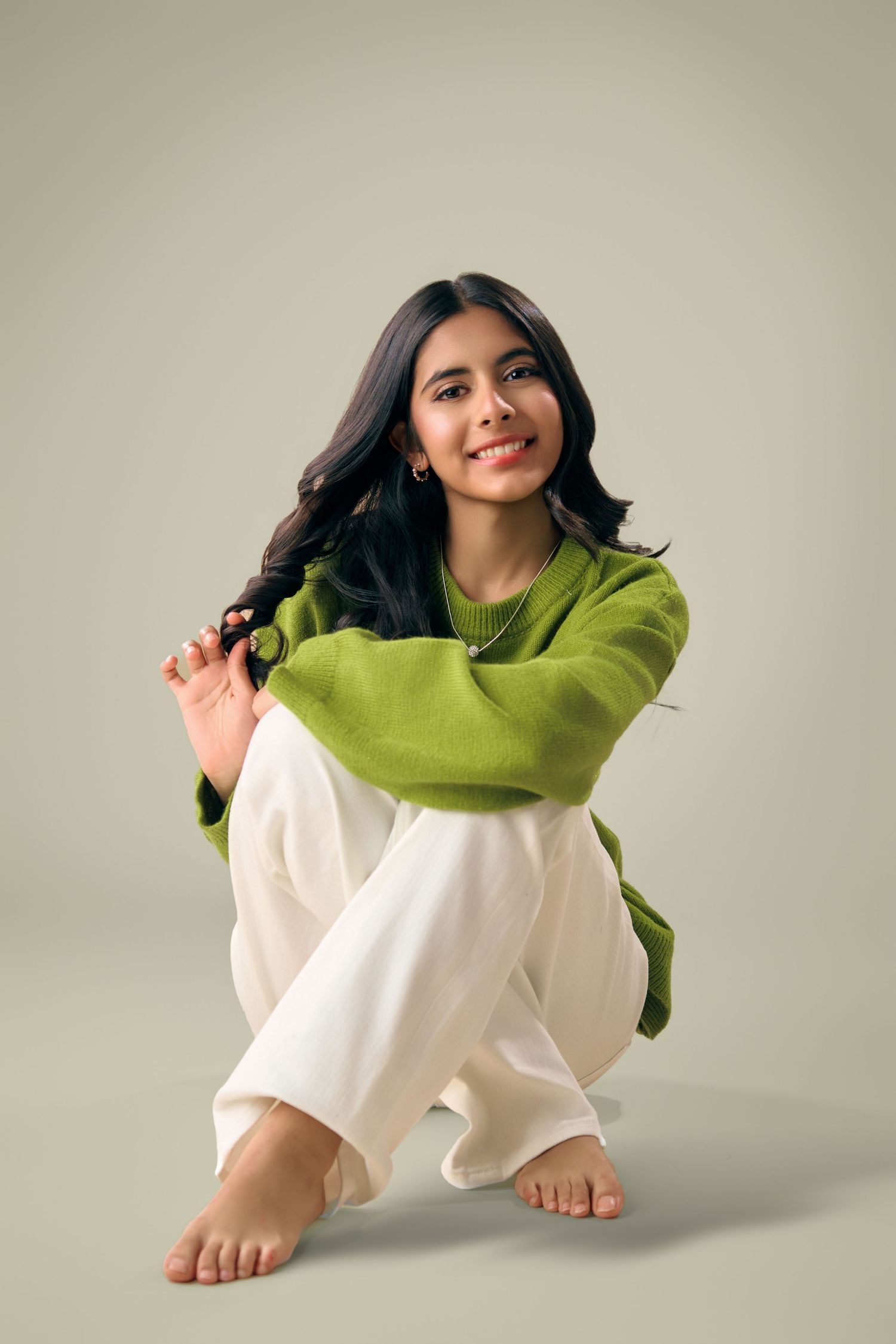 Smiling young woman in green sweater and white pants sitting barefoot on gray background.