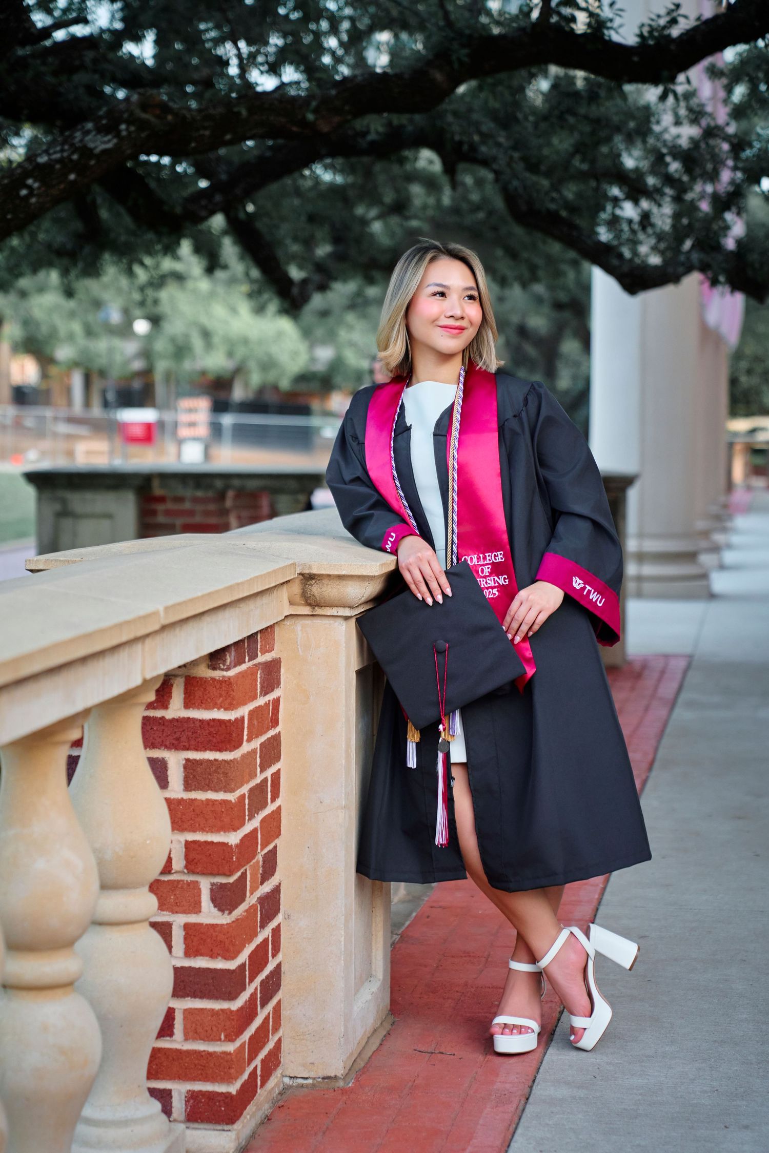 Female graduate in black cap and gown with pink TWU stole posing on campus steps.