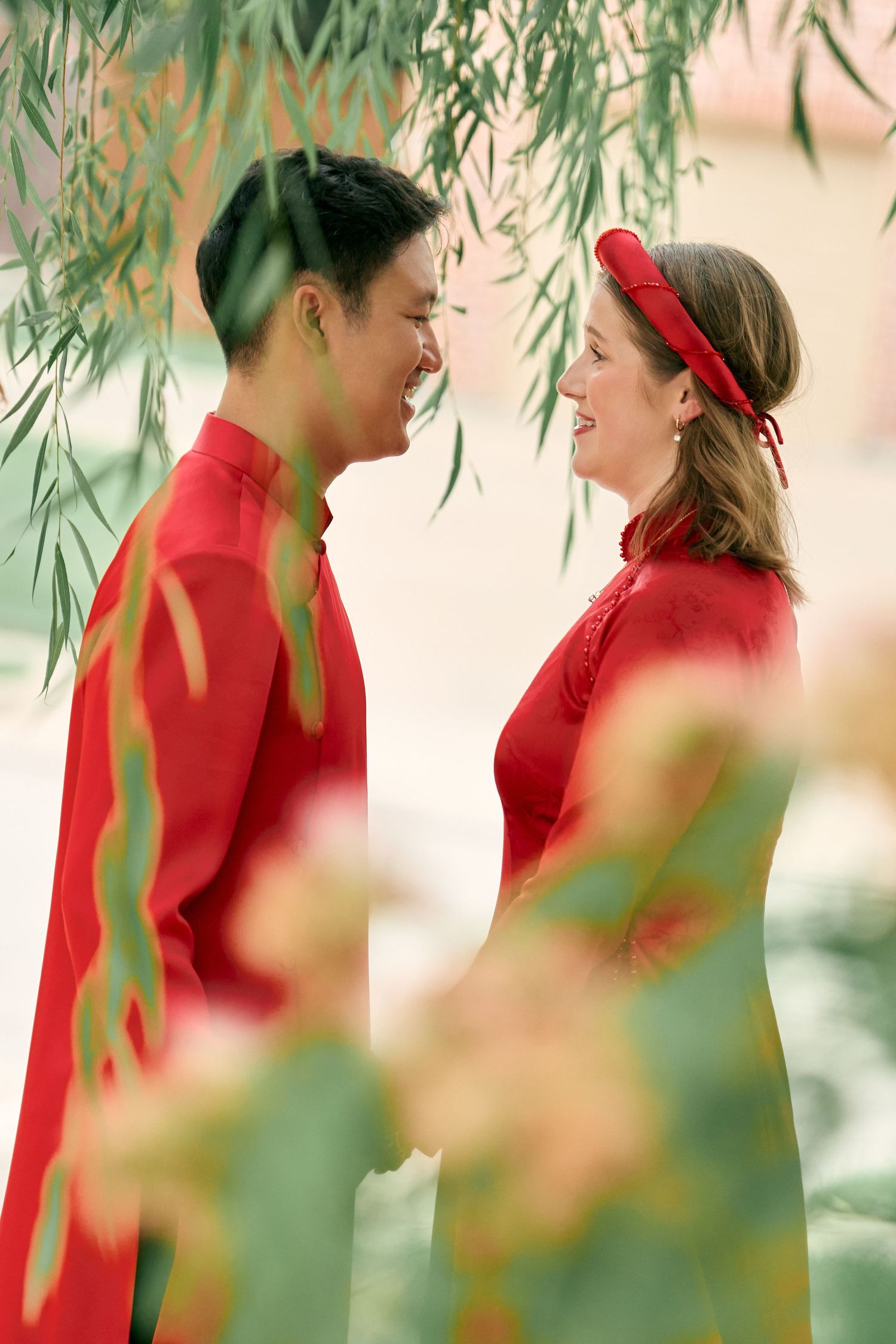 Couple in matching red traditional Vietnamese ao dai outfits smiling at each other under willow tree branches.