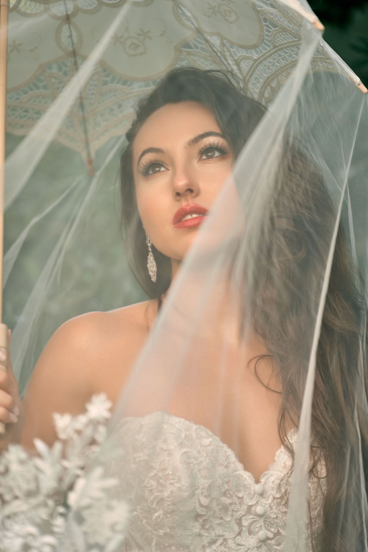 Elegant bride in lace wedding dress holding a sheer veil and floral bouquet, wearing crystal earrings.