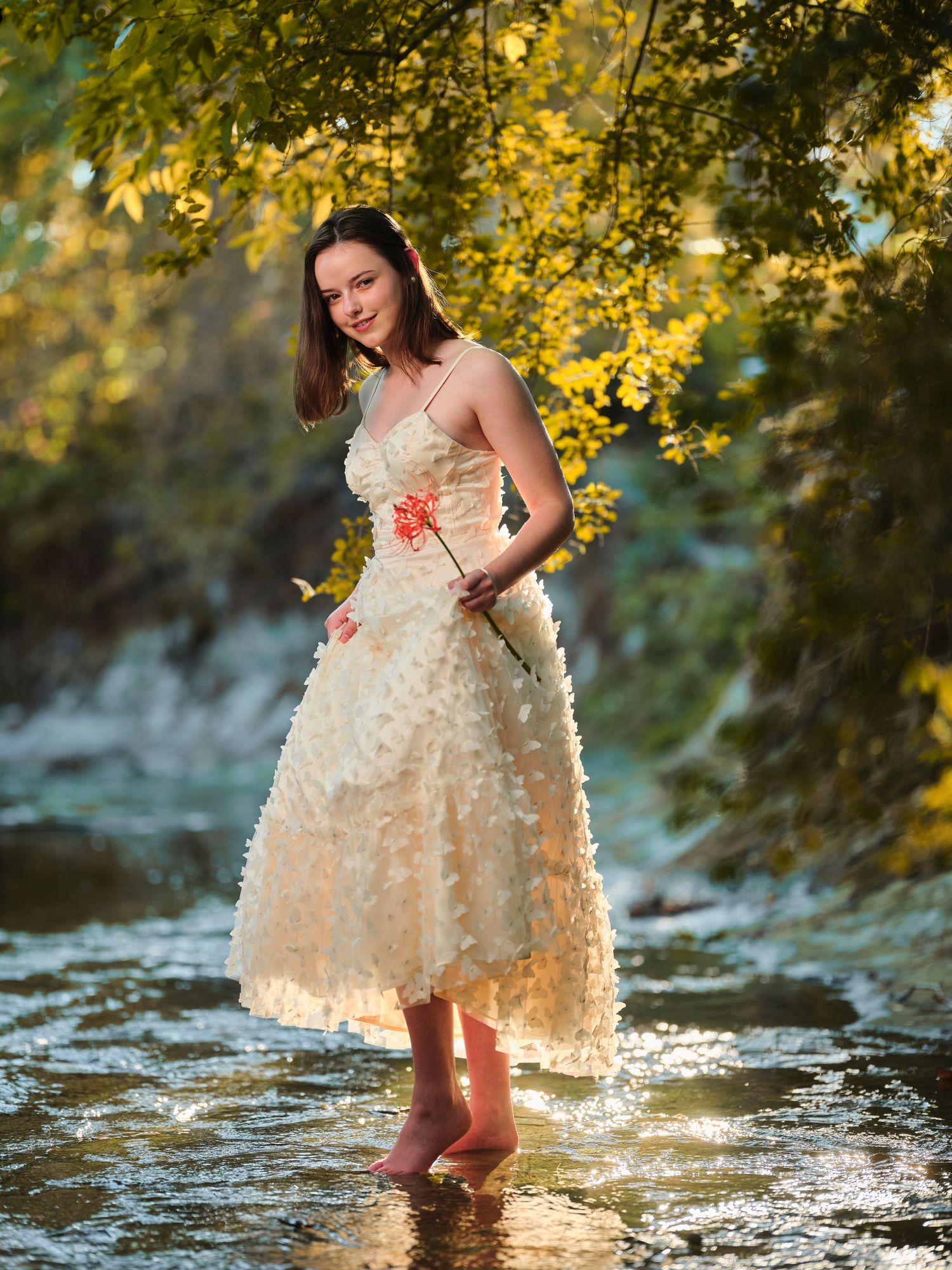 Young woman in cream ruffled dress standing barefoot in a sunlit stream surrounded by autumn foliage.