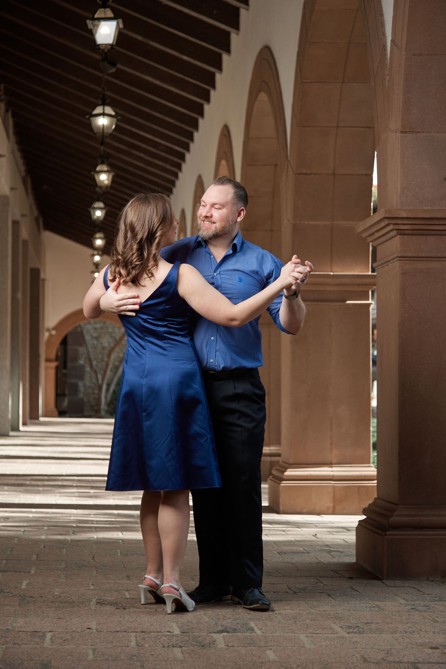 Elegant couple dancing tango in blue outfits under arched colonnade with warm lighting.