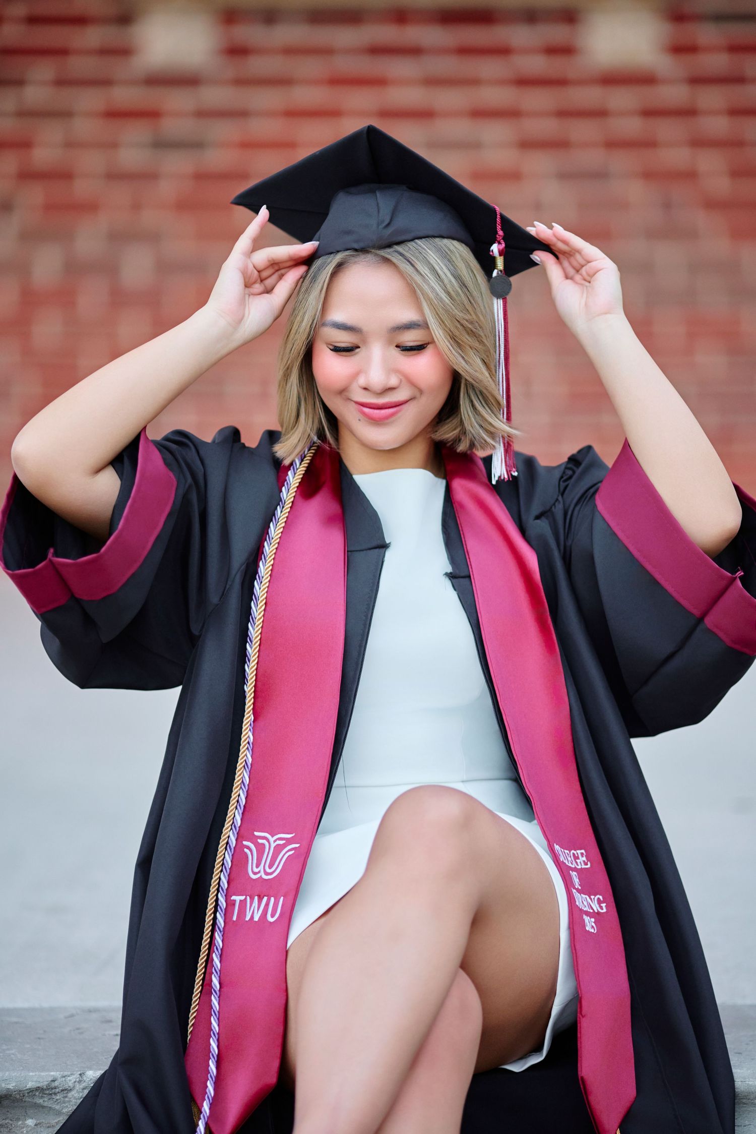 Smiling graduate adjusts black cap wearing TWU maroon graduation regalia.