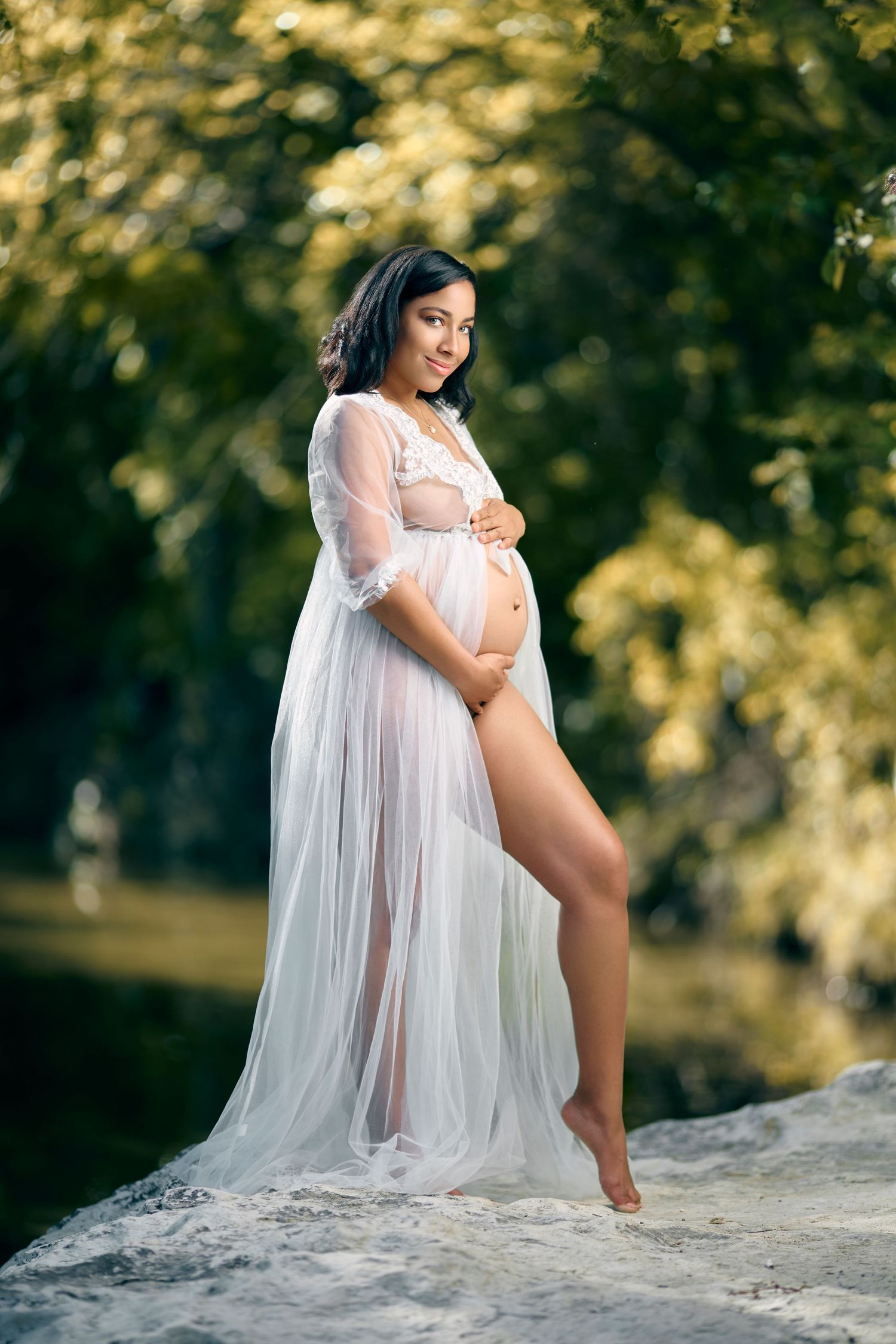 Pregnant woman in sheer white lace robe posing outdoors on rocks near a river.