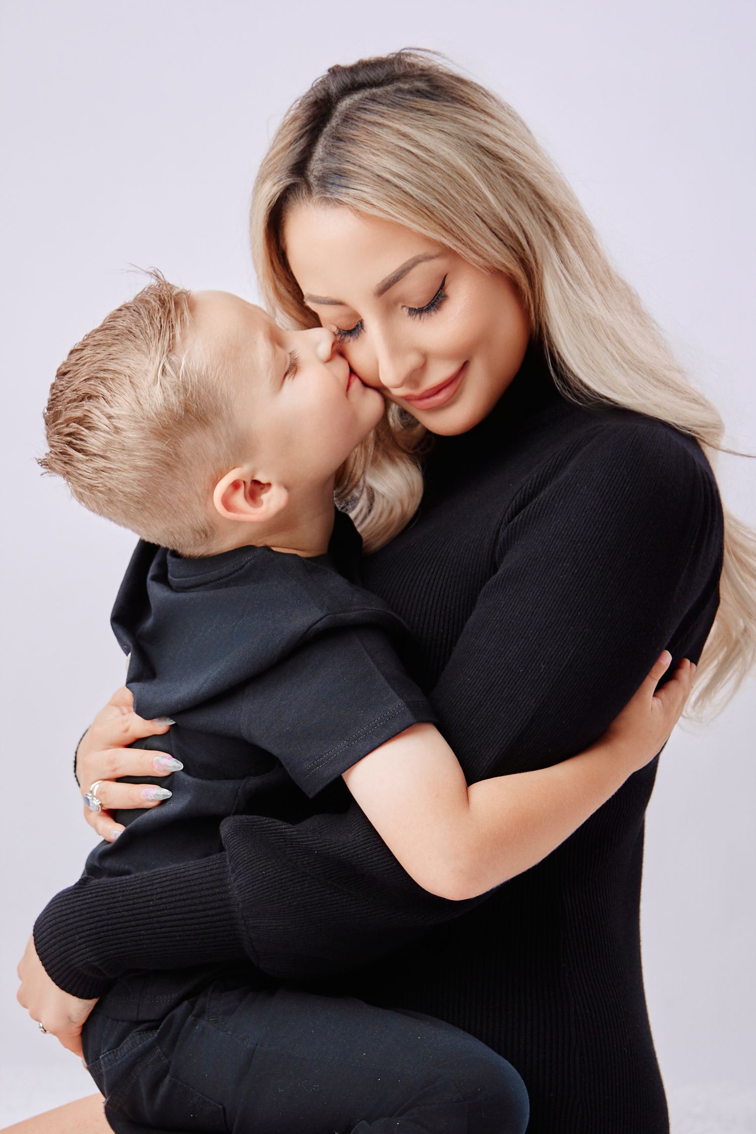 Blonde woman in black outfit holding young son who gives her a kiss on the cheek in studio.