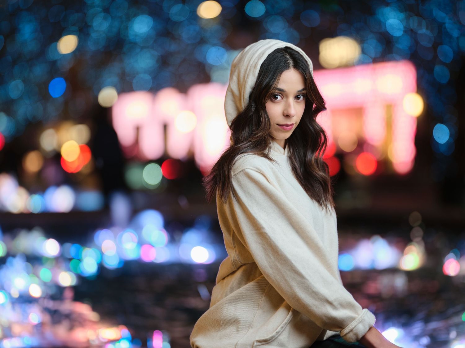 Young woman in beige hoodie posing outdoors at night with colorful bokeh city lights behind her.