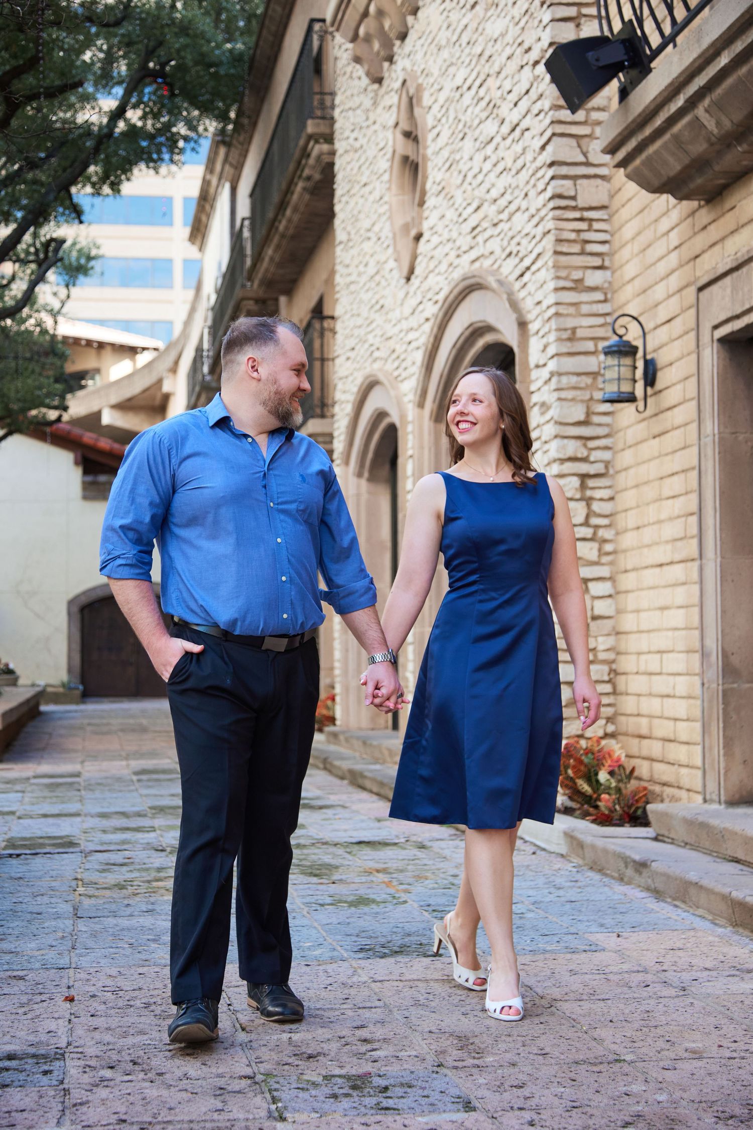 Happy couple holding hands walking together near historic stone building in matching blue outfits.