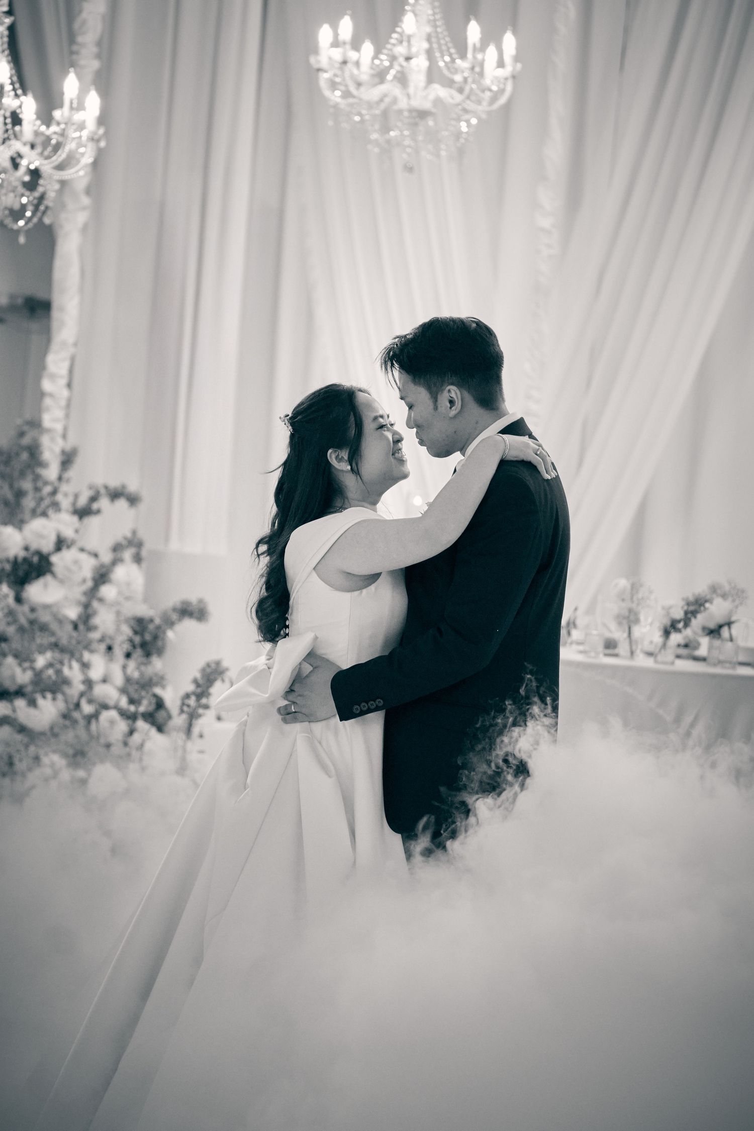 Black and white photo of a bride and groom sharing a romantic embrace on a misty dance floor beneath a crystal chandelier.