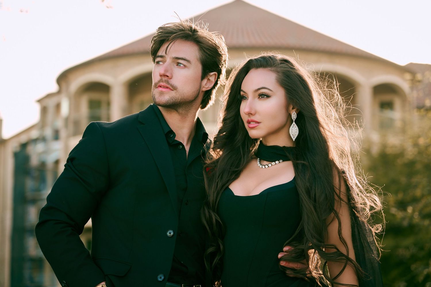 A stylish couple in black outfits poses outdoors at golden hour, with a woman in a choker and a man in a blazer.