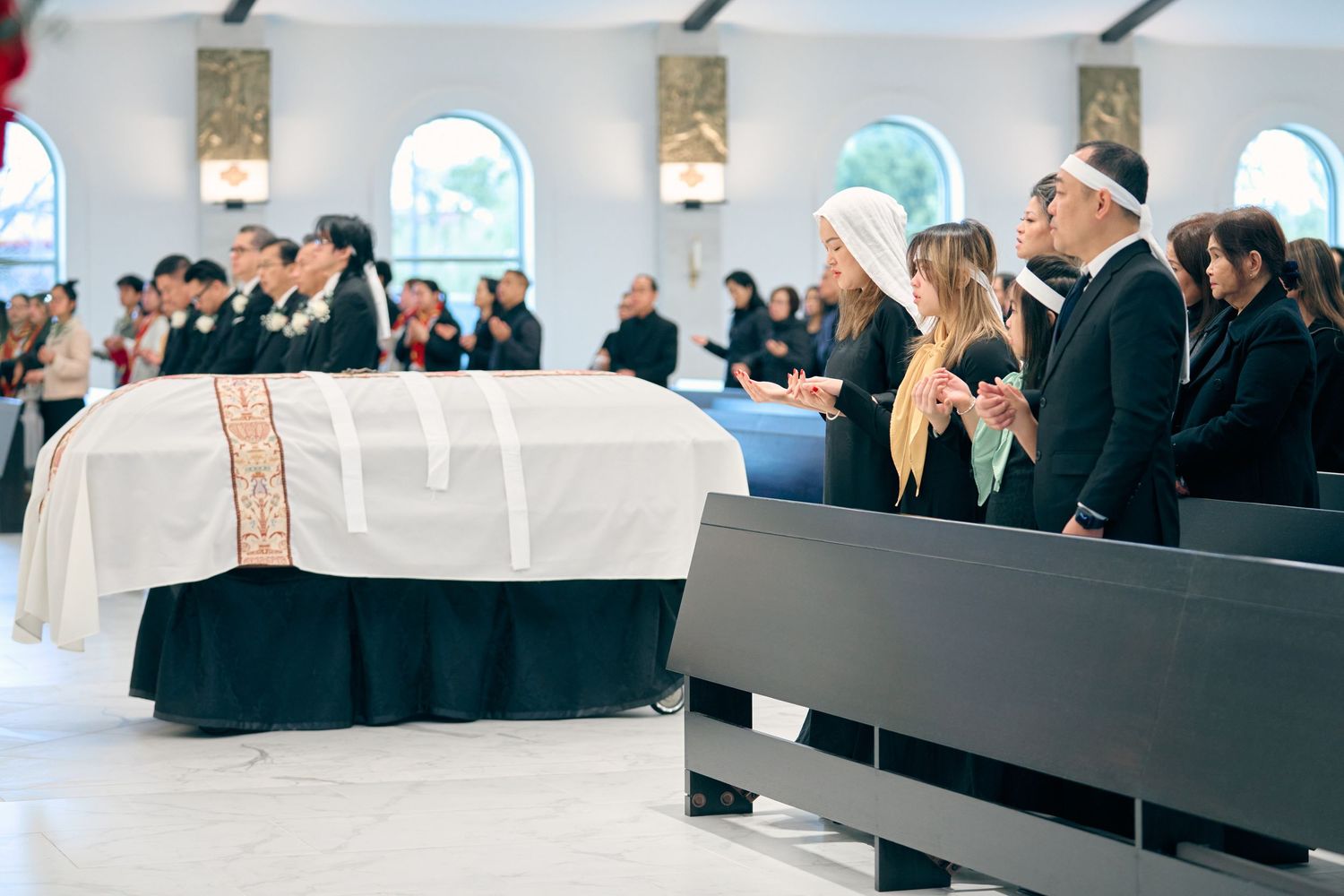 Catholic funeral Mass with casket draped in white pall, mourners standing in prayer inside a modern church.