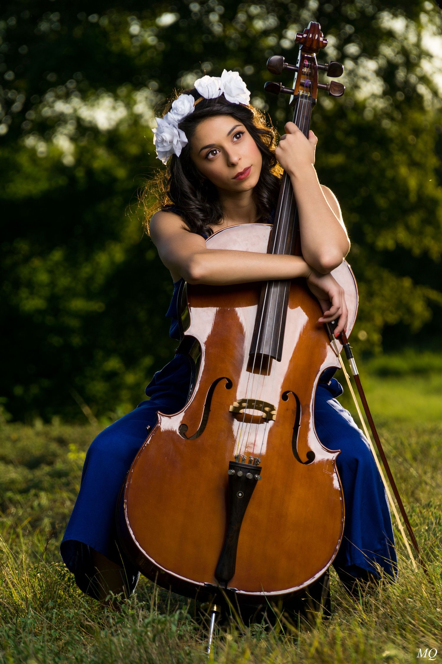 Young woman with white flower crown embracing a cello while sitting in a sunlit outdoor meadow.