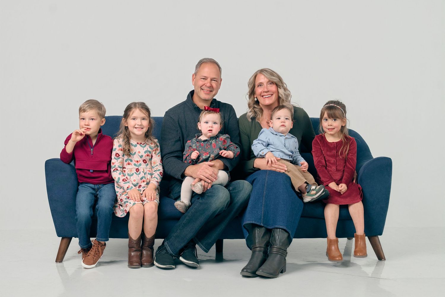 A family of seven with five young children poses together on a blue couch in a studio portrait.