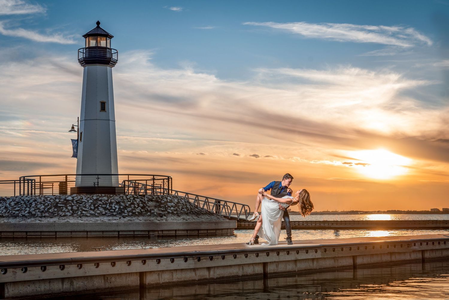 Romantic couple dancing near lighthouse at stunning golden sunset over calm harbor waters.