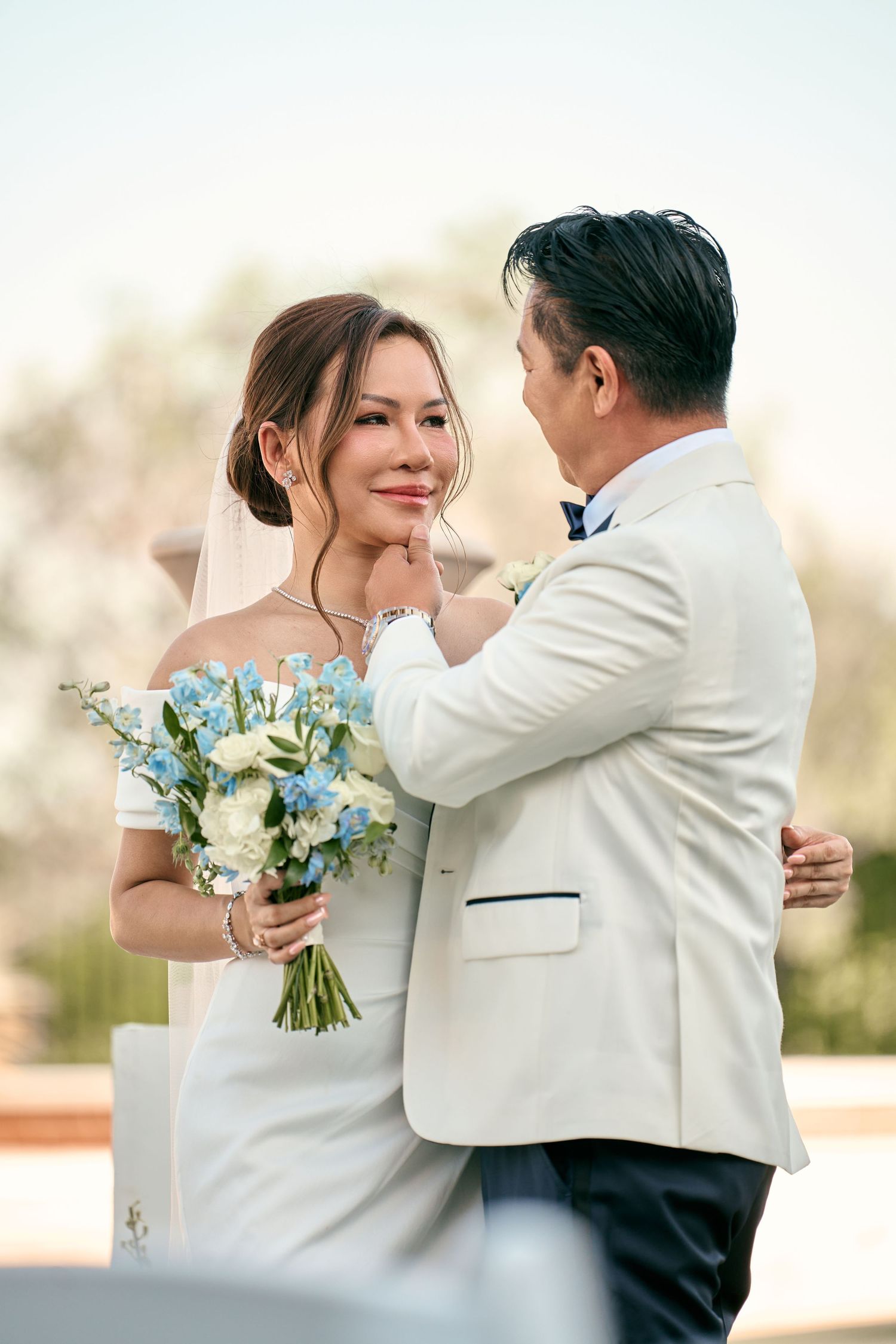 A bride and groom share a tender moment outdoors, with the groom gently touching the bride's chin as she holds a blue and white bouquet.