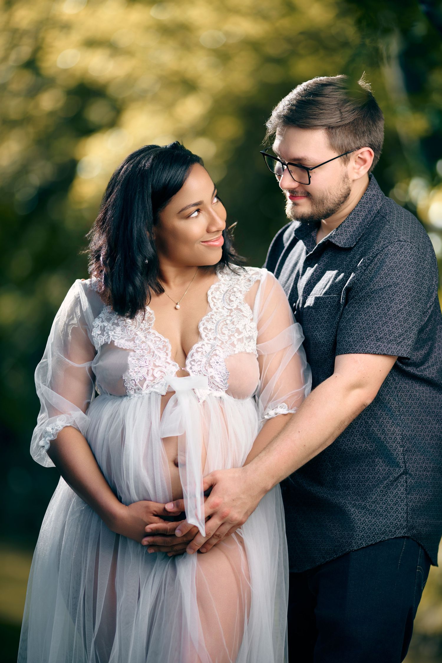Happy expectant couple in park, woman in sheer white lace maternity gown, partner embracing her bump.