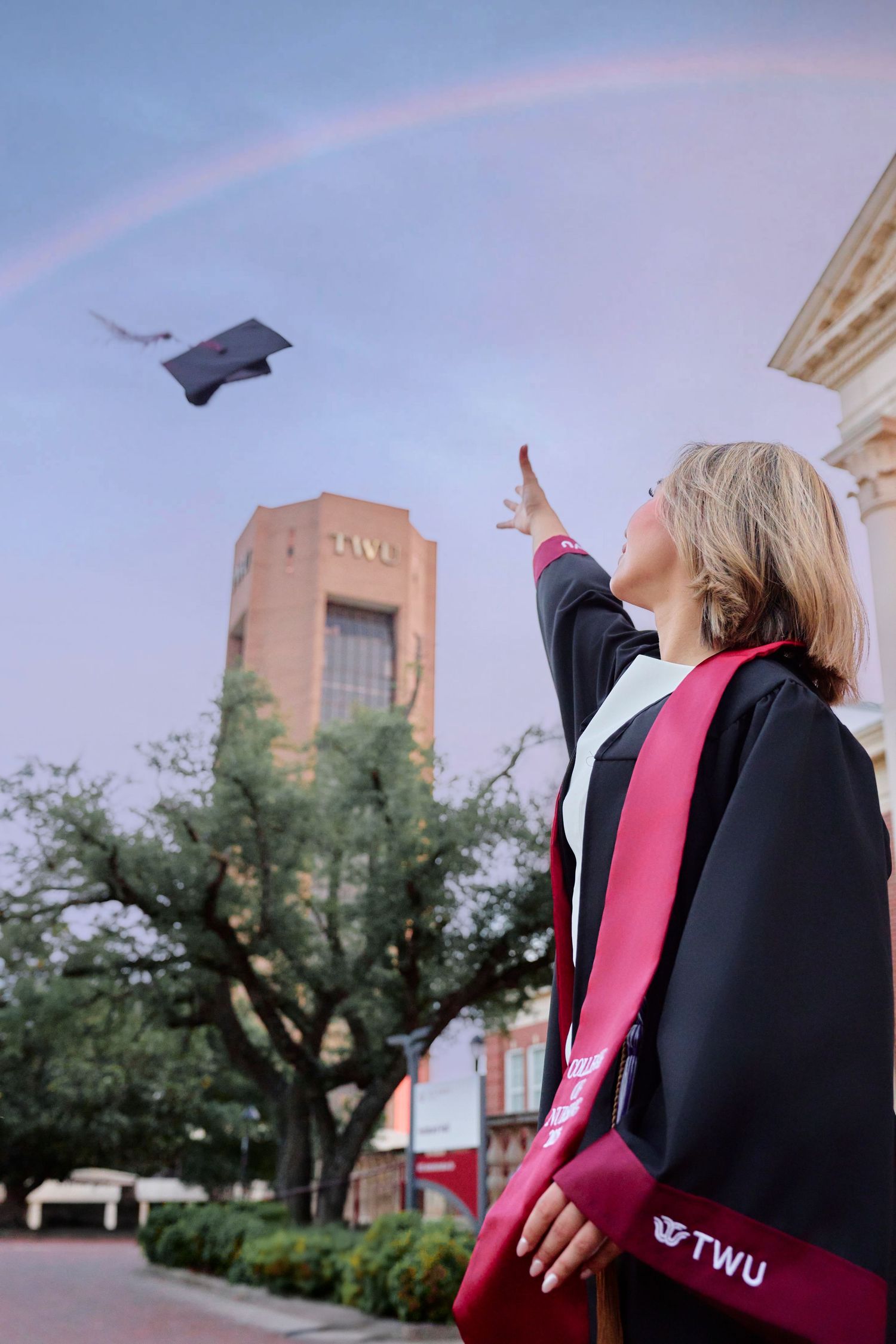TWU graduate tosses cap toward rainbow sky near university tower building.