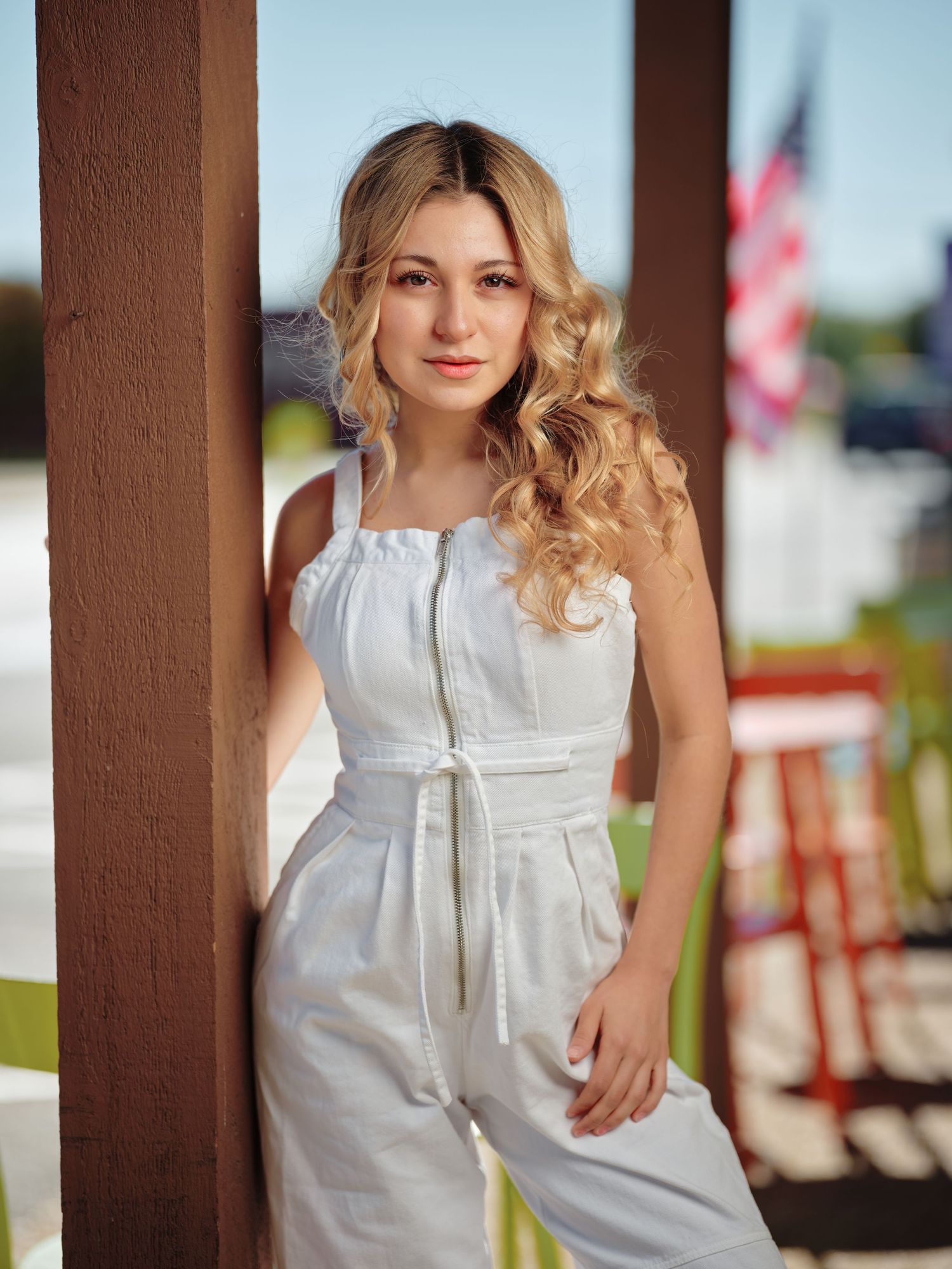 Young blonde woman in a white jumpsuit posing by a wooden post with an American flag in the background.