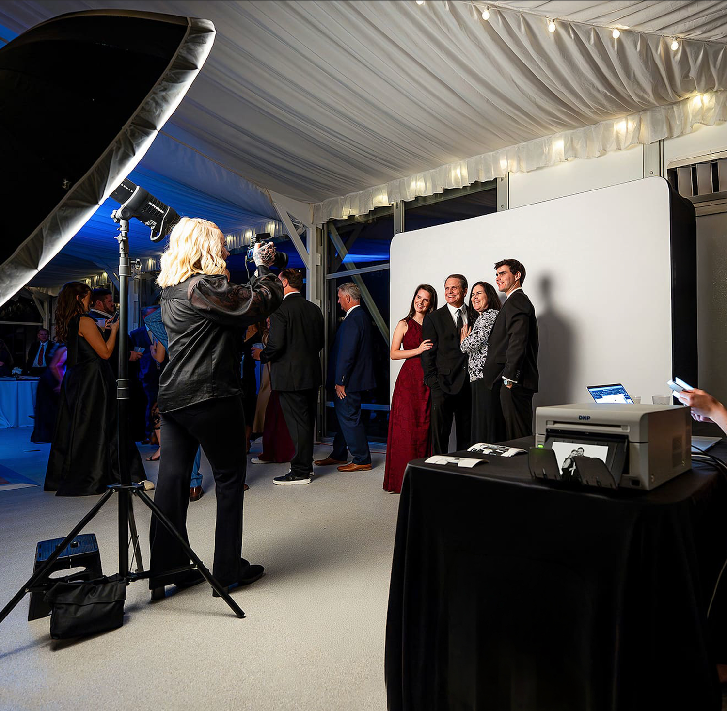 A photographer captures a group portrait at a formal event inside a tent with professional lighting equipment.