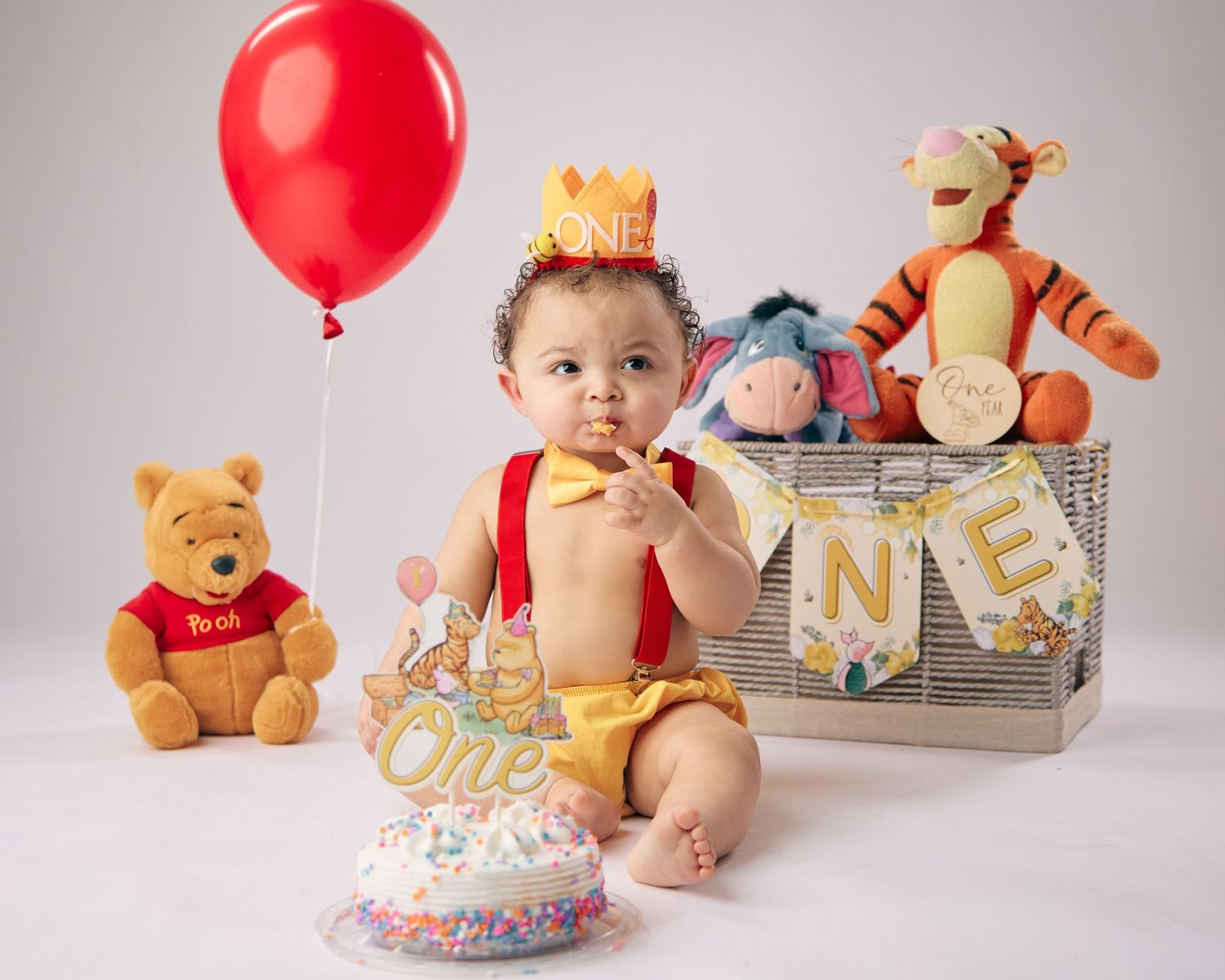Baby's first birthday cake smash with Winnie the Pooh theme, red balloon, stuffed animals, and gold crown.