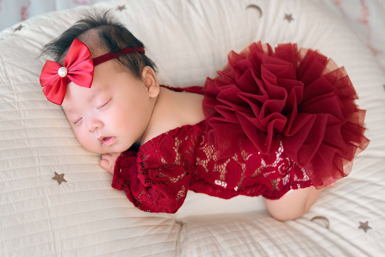 Sleeping newborn baby in red lace outfit and bow headband on white pillow with star decorations.