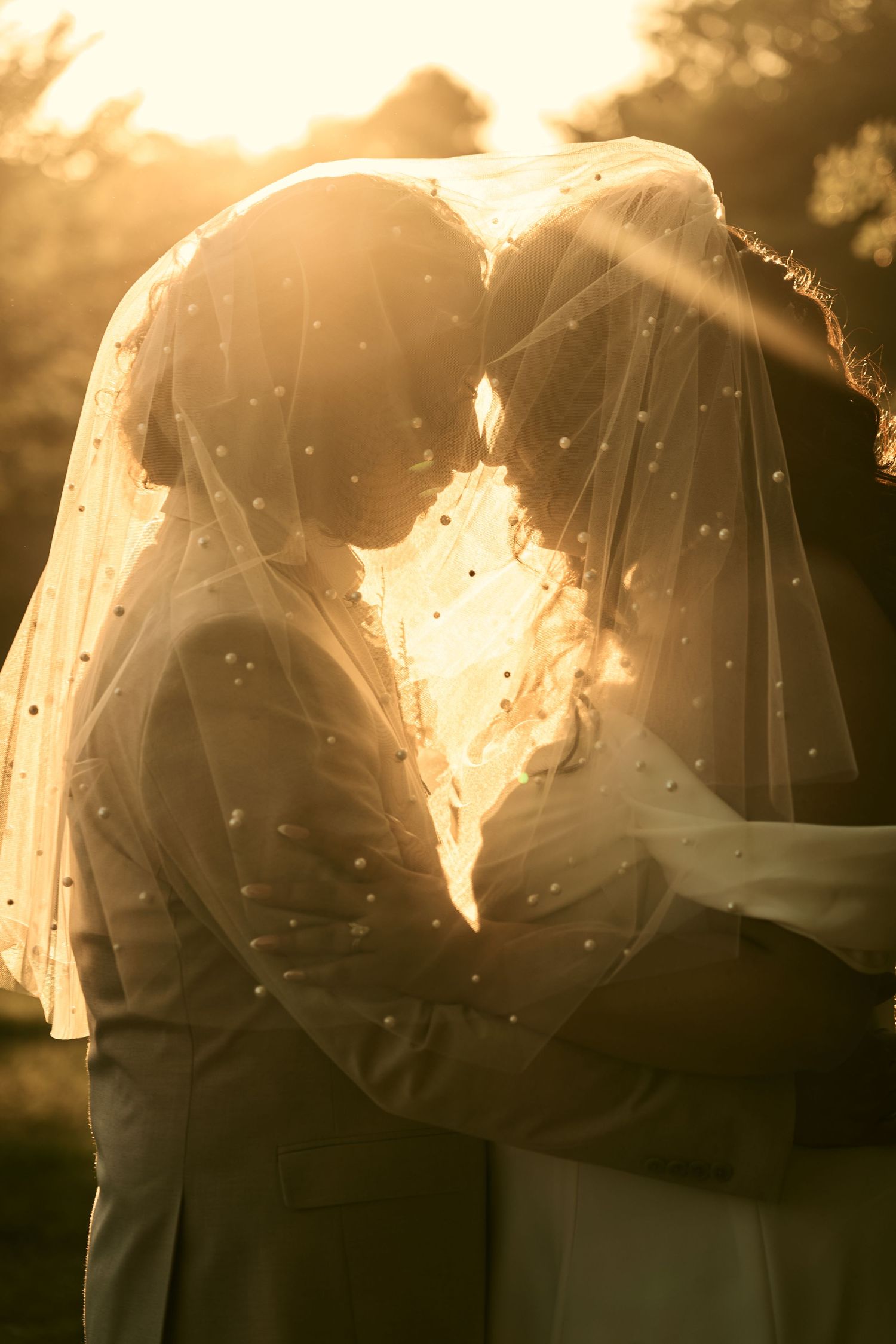 Bride and groom sharing a romantic moment under a pearl-dotted veil, backlit by golden sunset light.