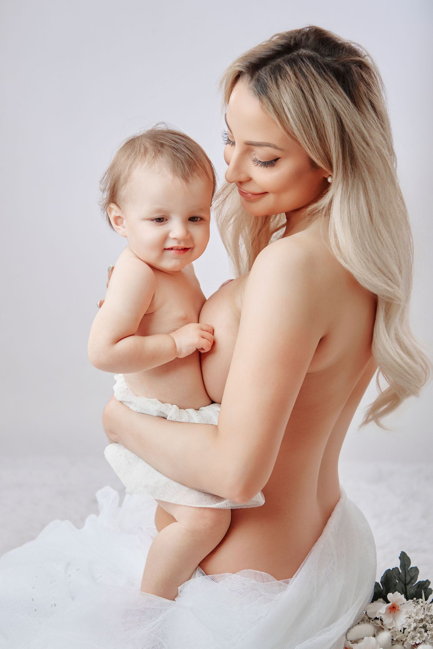 A blonde mother tenderly holds her smiling baby wrapped in white fabric during a soft studio portrait session.