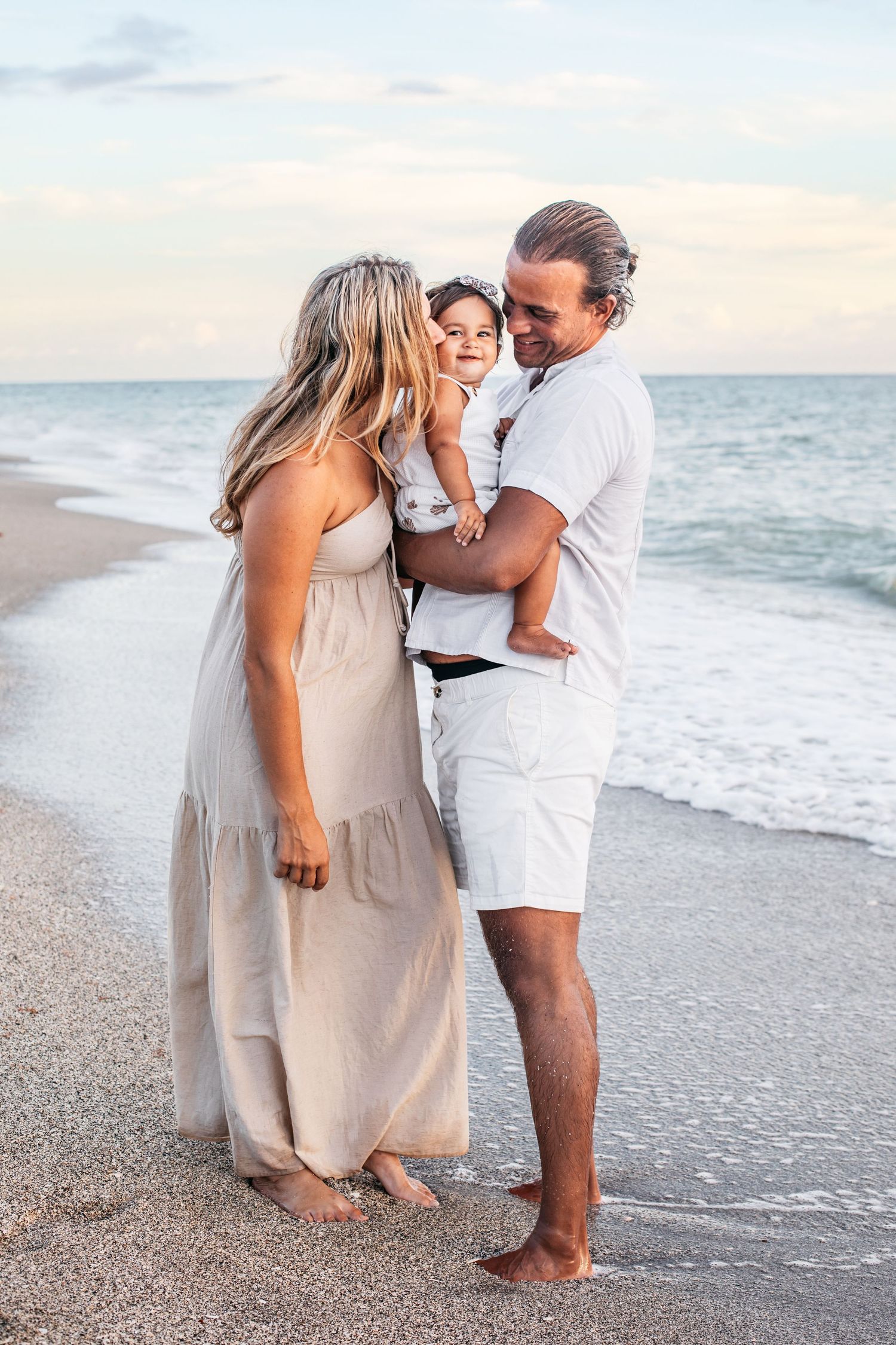 A couple shares a tender moment on the beach at sunset wearing coordinating beige and white outfits.