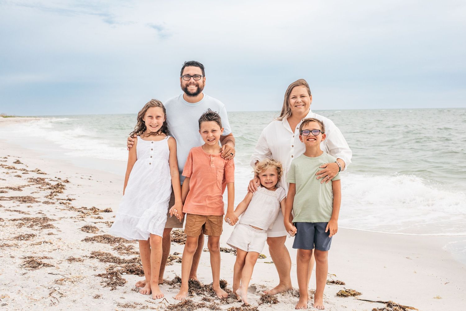 Smiling family poses together on a sandy beach with the ocean and cloudy sky in the background.