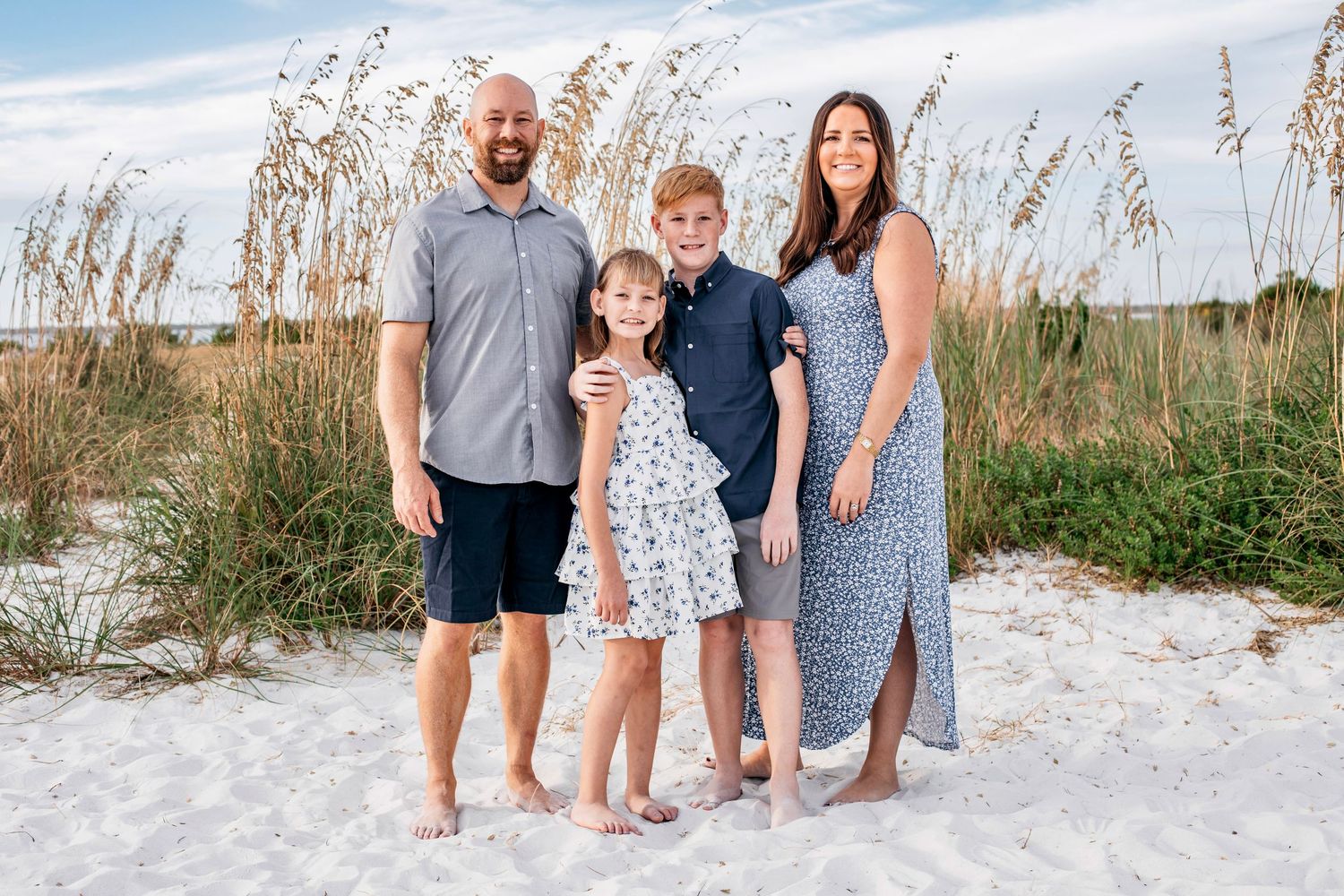 A group of four people stand together smiling on a white sandy beach with sea oats in the background.