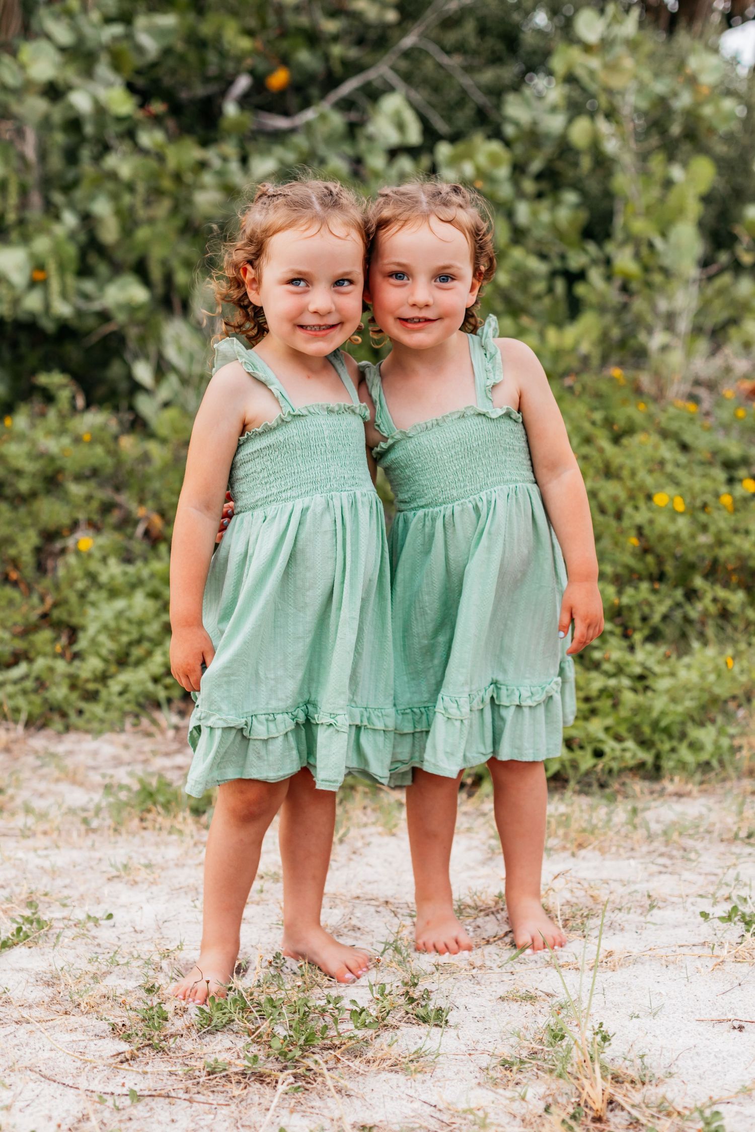 Two young twin girls in matching mint green smocked dresses stand barefoot on sand with green foliage behind them.