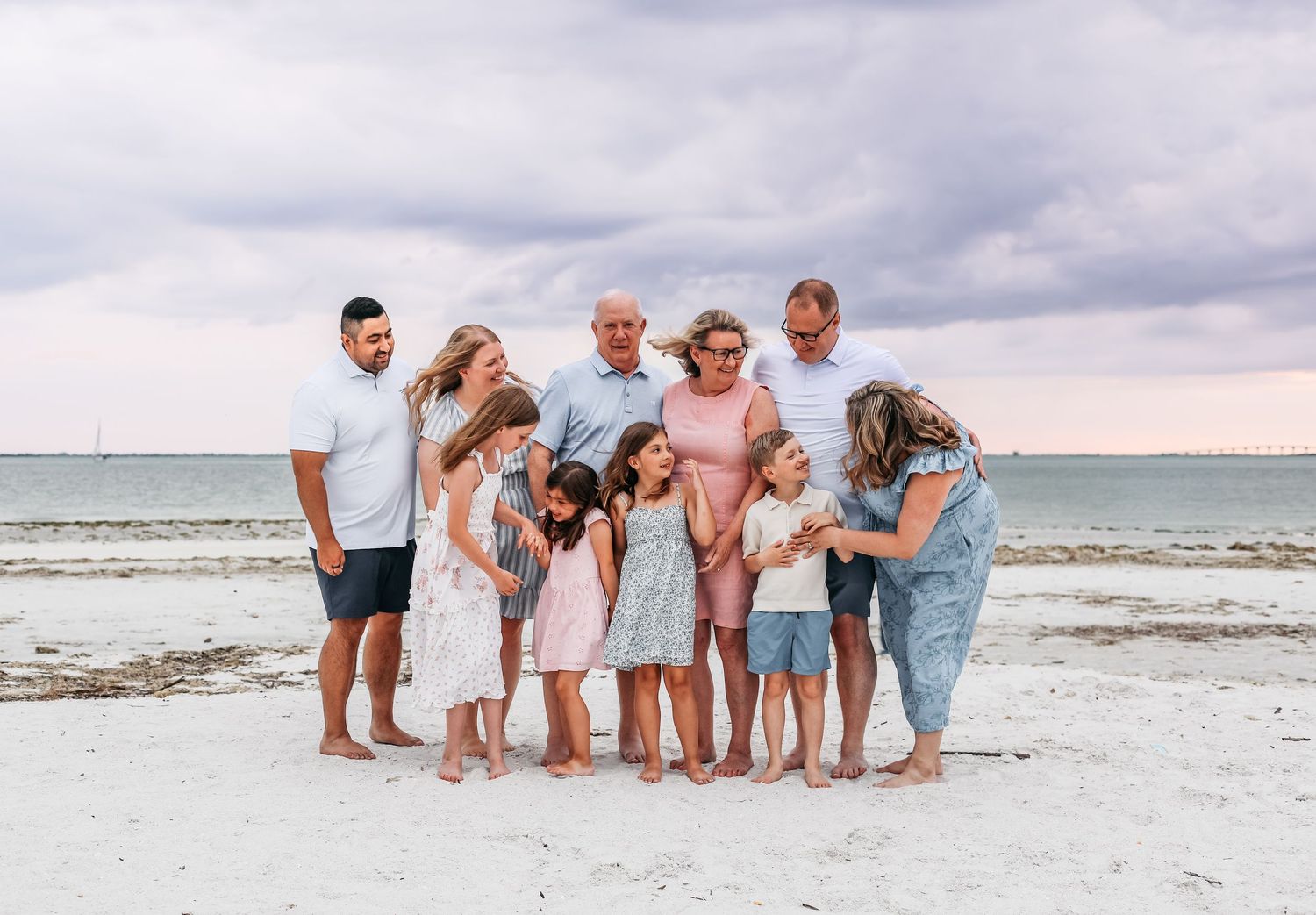 Large family group photo on white sand beach with cloudy sky, adults and children smiling together near the ocean.