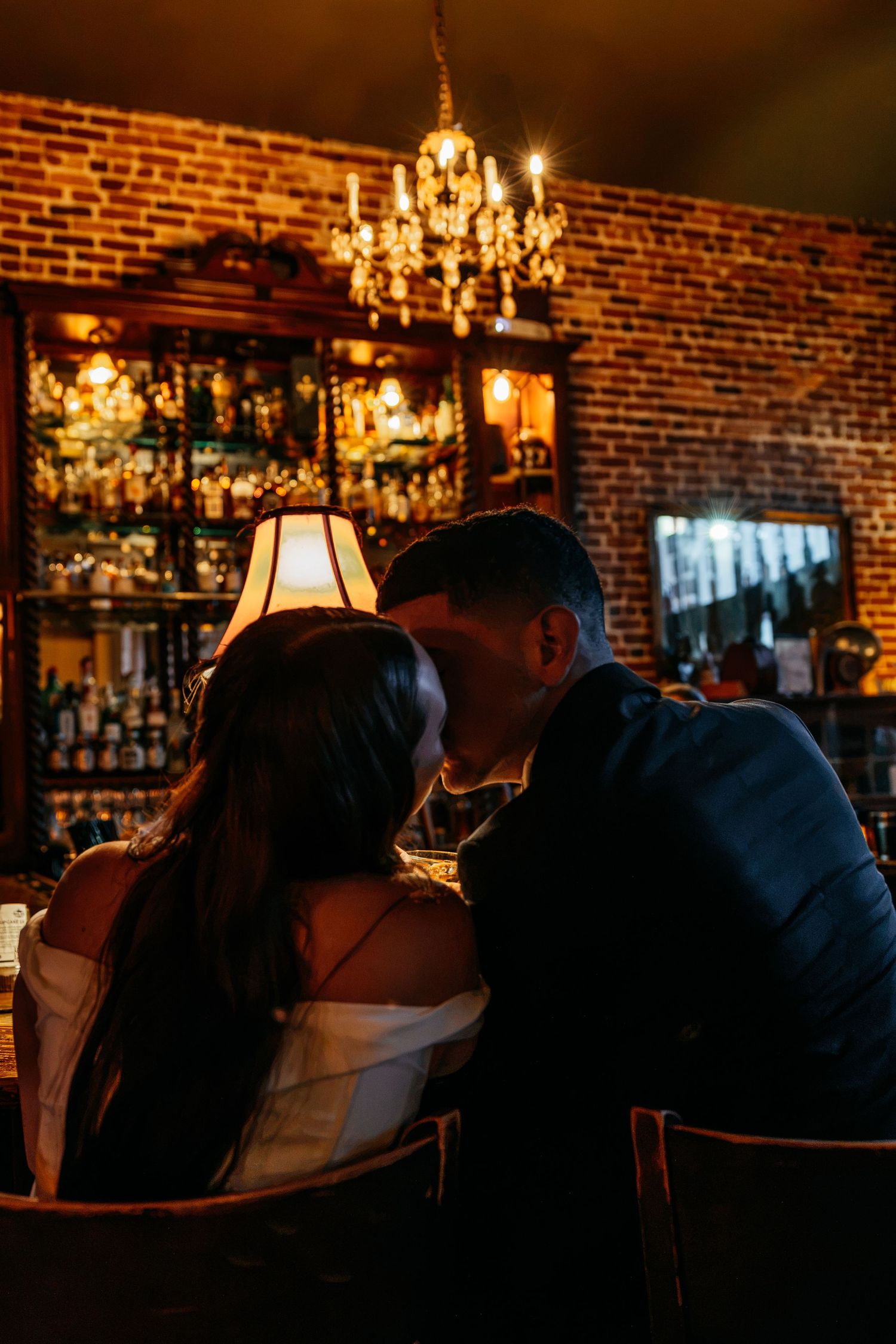 Couple sharing a romantic kiss at a dimly lit bar with warm chandelier lighting and exposed brick walls.