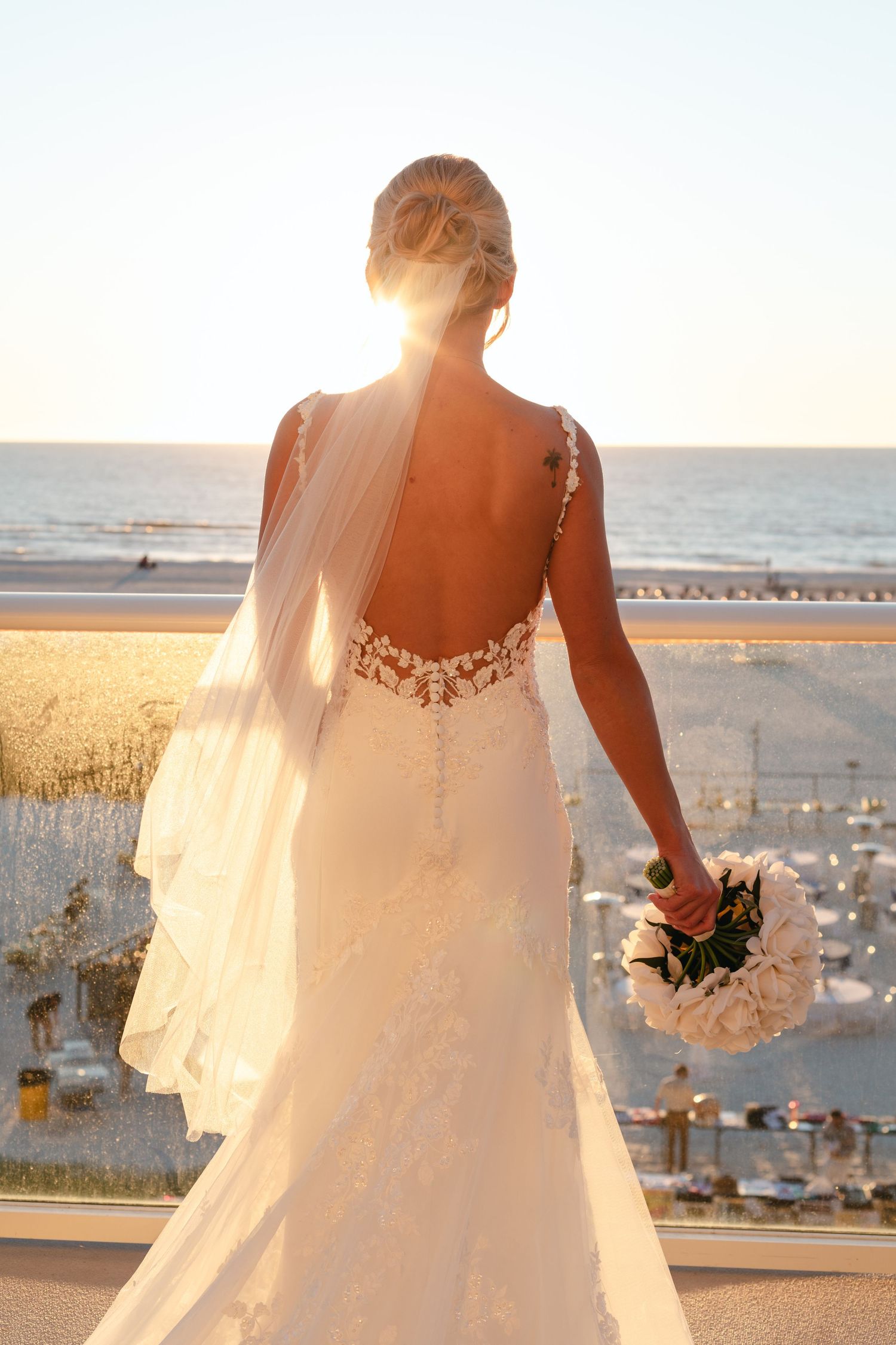 Bride in backless lace wedding gown holding bouquet, overlooking beach at sunset with veil flowing.